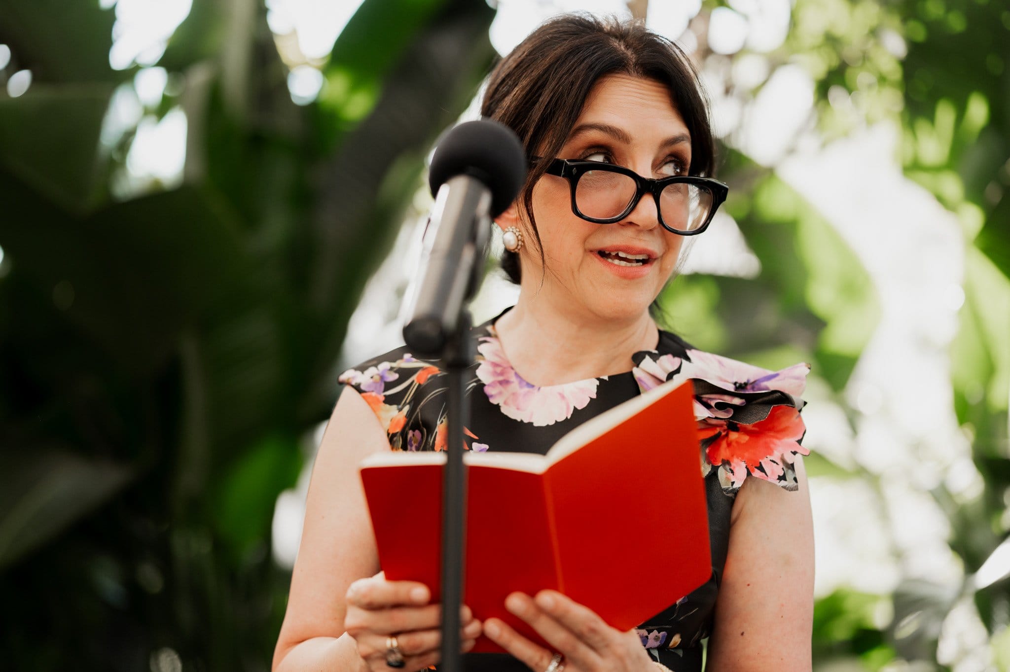 Standing near a microphone, a woman in floral attire reads from a red book, framed by lush greenery. The scene captures the essence of wedding photos at Blockhouse, with nature providing a perfect backdrop for the memorable moment.