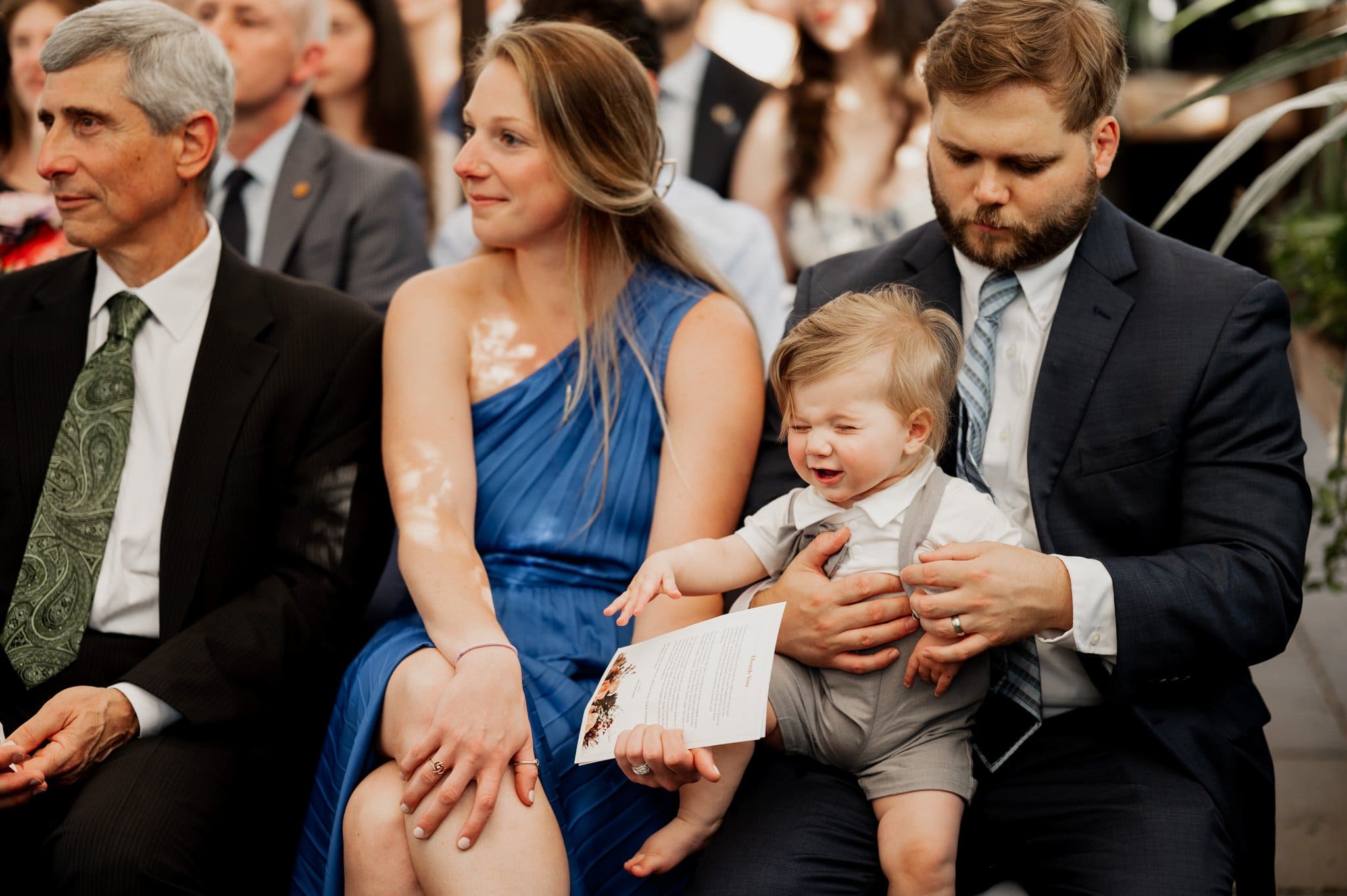 The moment captures three adults and a baby: a man cradling the child, a woman in a blue dress seated beside them, and another man in a dark suit. This charming scene is perfect for wedding photos at Blockhouse.