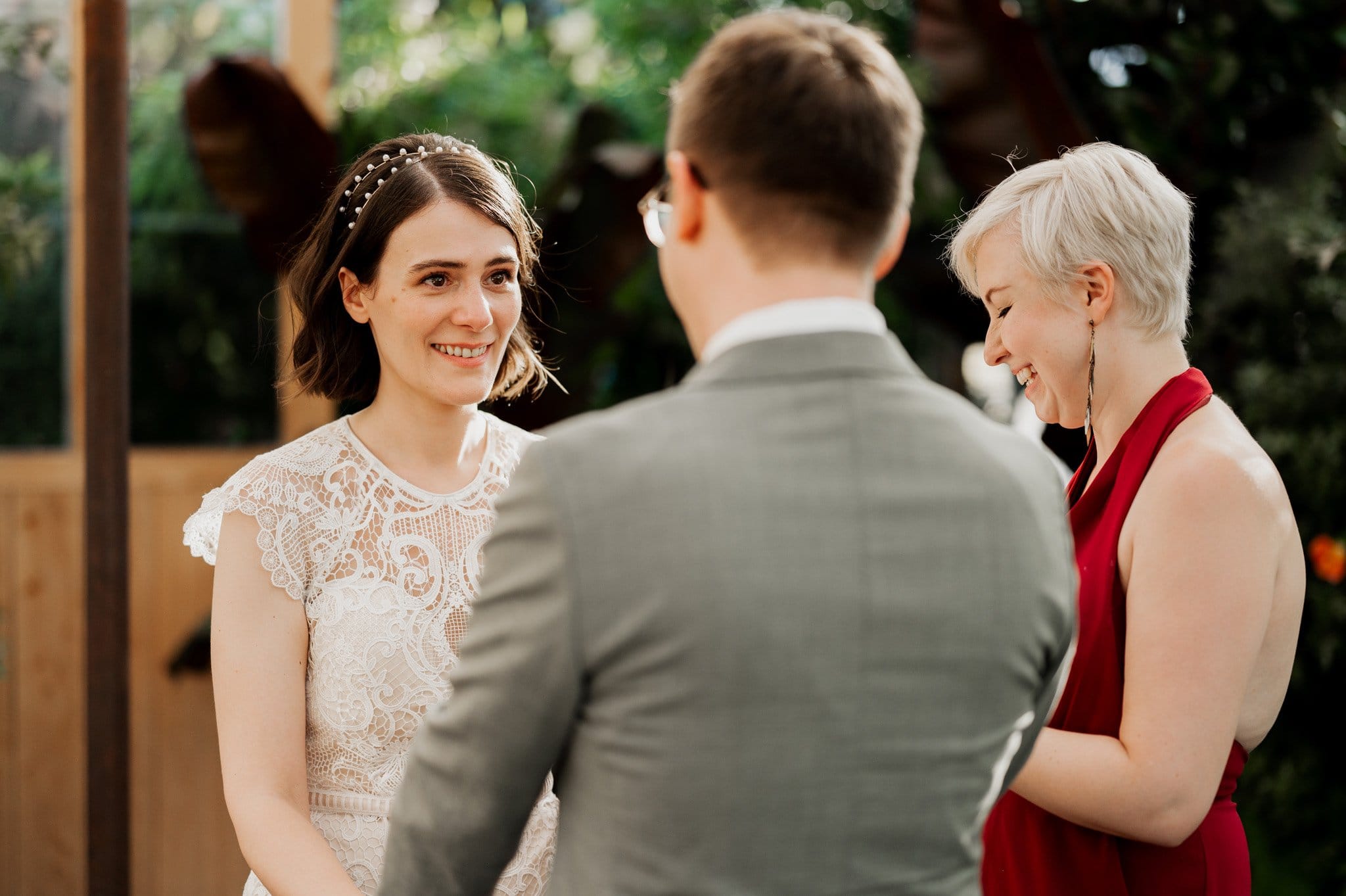 A couple exchanging vows outdoors, with the woman smiling and the man in a gray suit, as an officiant stands beside them. Theyre captured in stunning wedding photos at Blockhouse.
