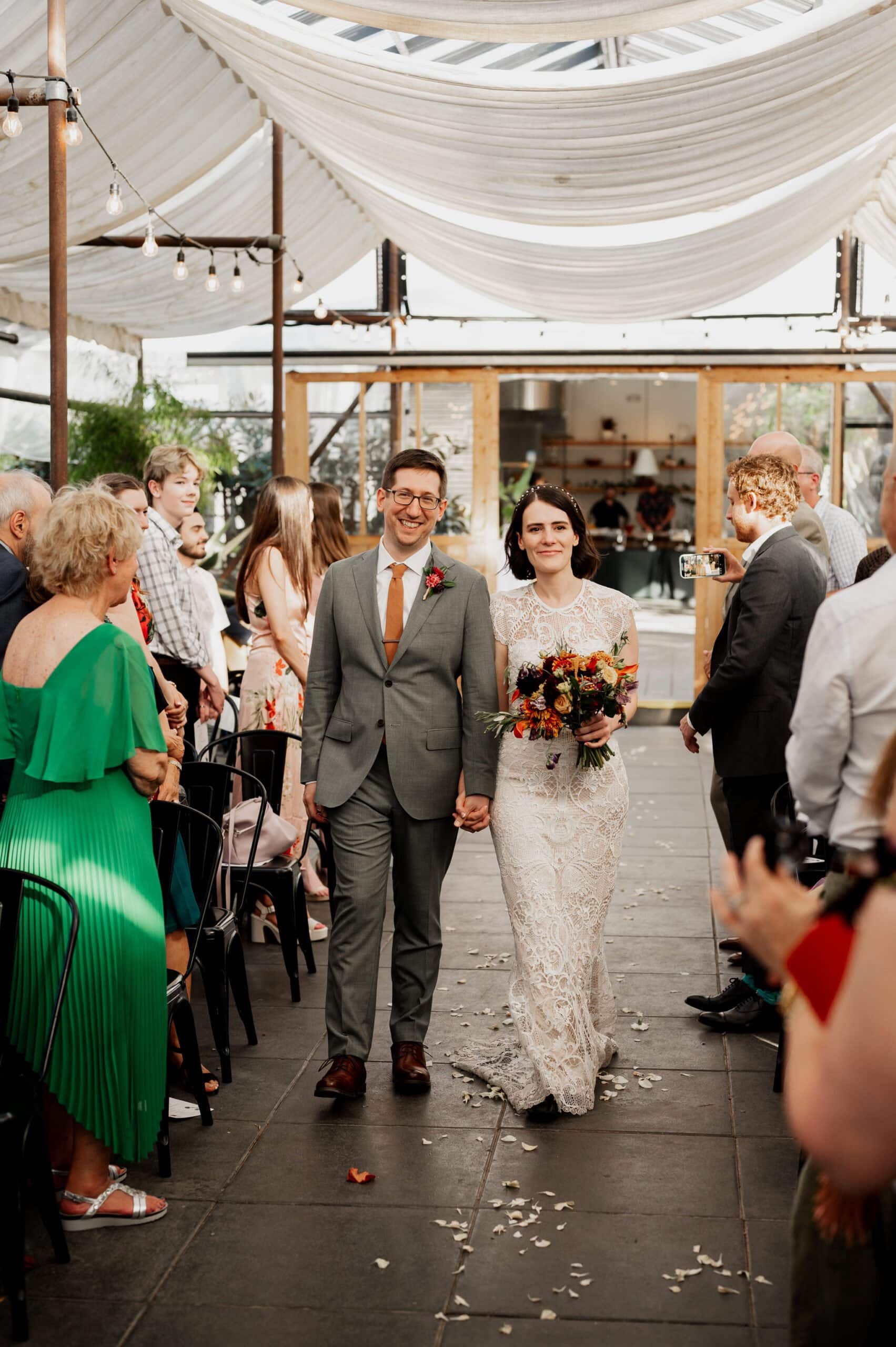 The bride and groom walk hand in hand down the aisle, surrounded by guests in an indoor venue adorned with white drapes above, perfect for capturing wedding photos at Blockhouse.