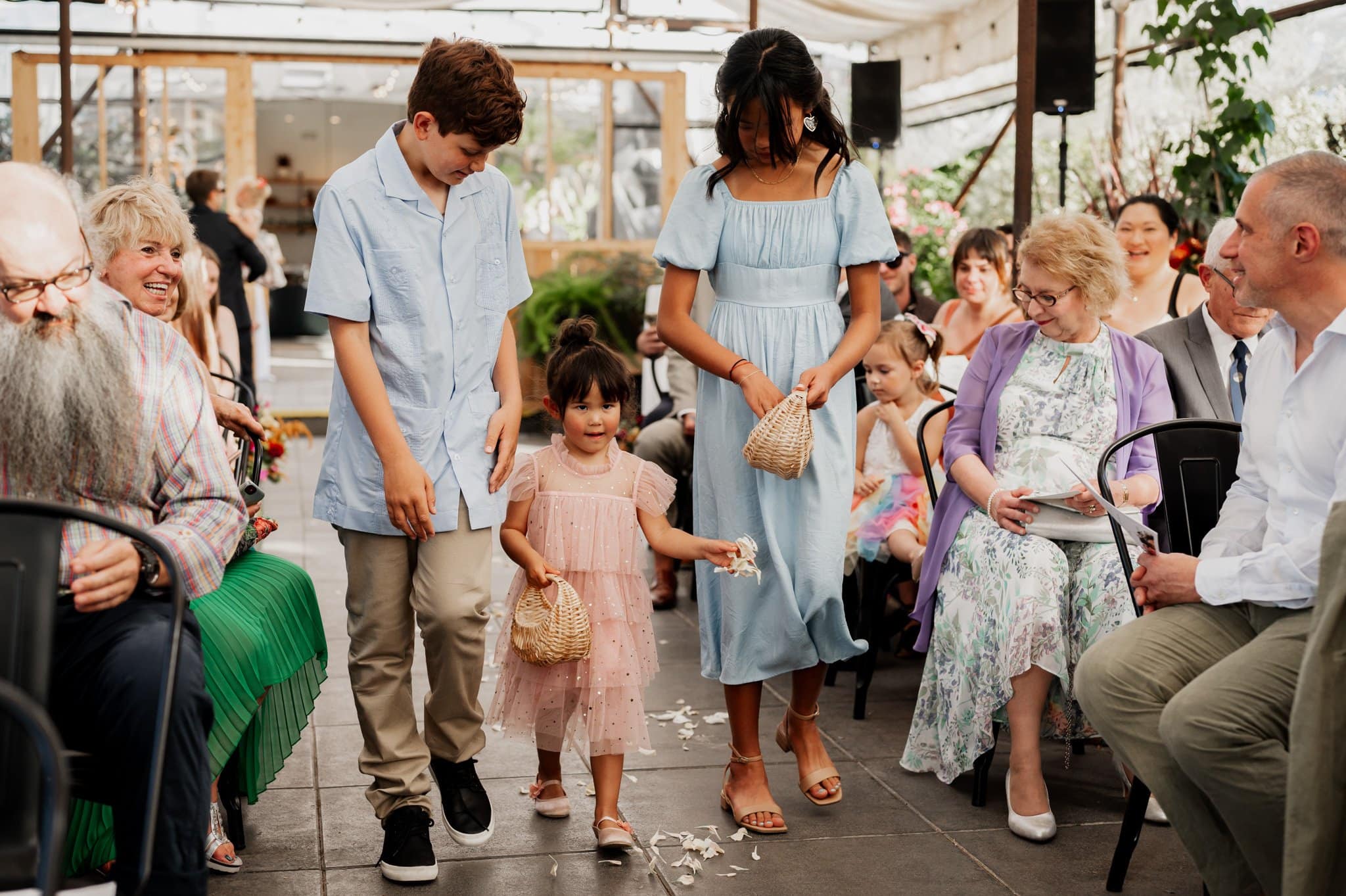 At an indoor event, three children walk down the aisle, scattering petals as guests smile and watch. Capture the magic akin to wedding photos at Blockhouse.