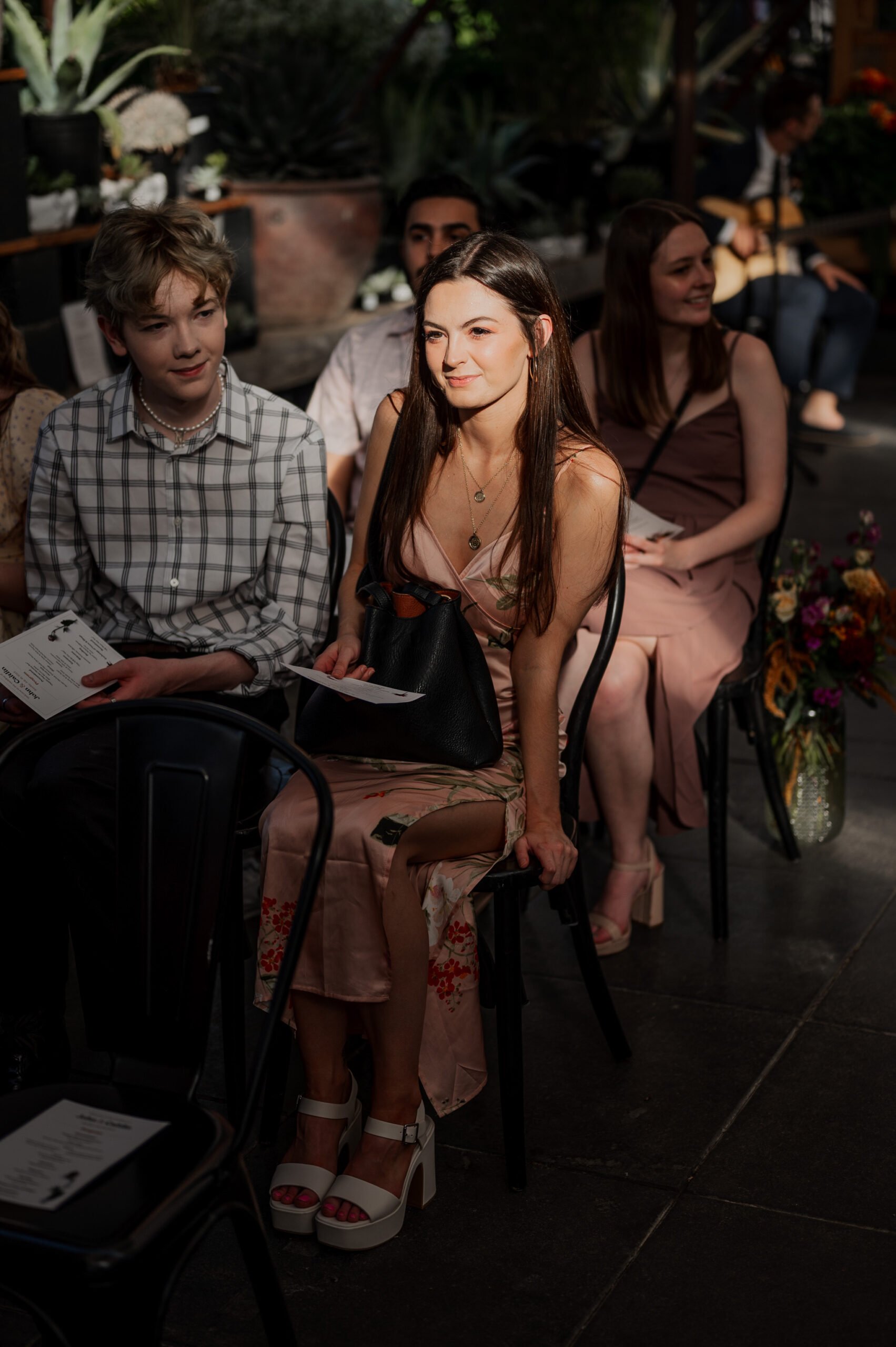A group is seated on chairs, dressed semi-formally, in a sunlit room adorned with plants. The setting captures the essence of wedding photos at Blockhouse, where natural light enhances the atmosphere.