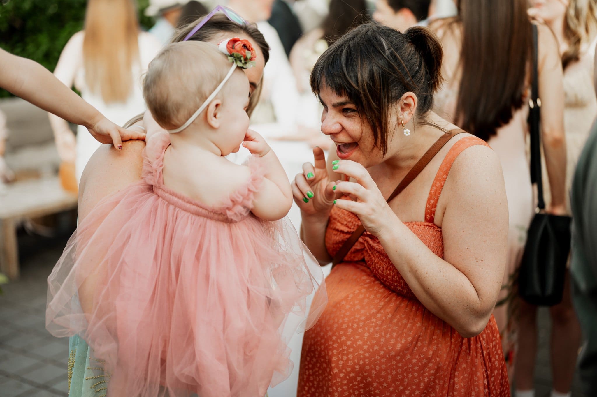 A woman joyfully interacts with a baby in a pink dress and headband at an outdoor gathering, creating moments perfect for wedding photos at Blockhouse.