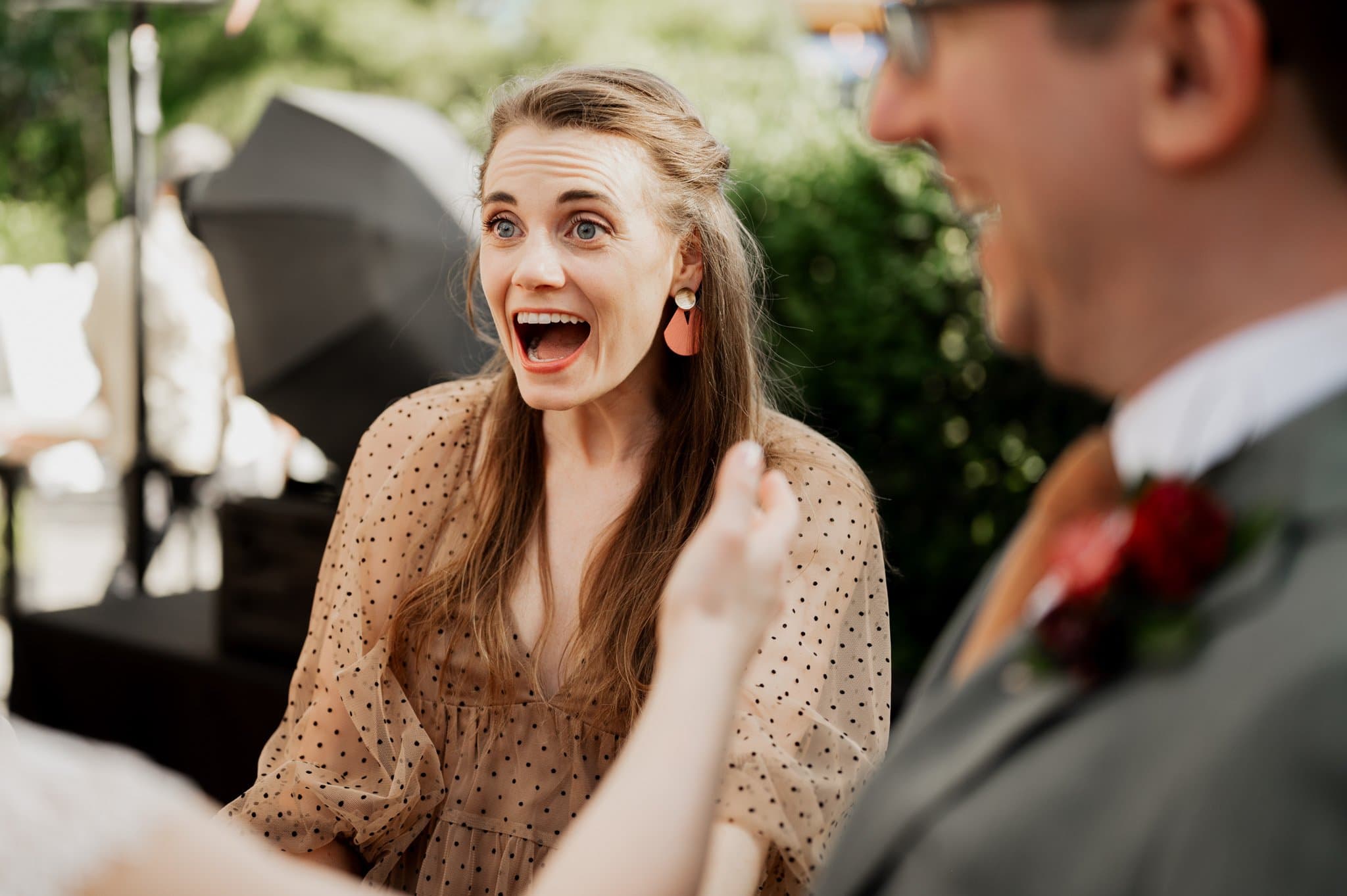 The woman with long hair in a polka dot dress looks surprised and happy outdoors, chatting with a man in a suit. Their laughter echoes through the garden, as if captured in wedding photos at Blockhouse, where everything seems beautifully spontaneous.