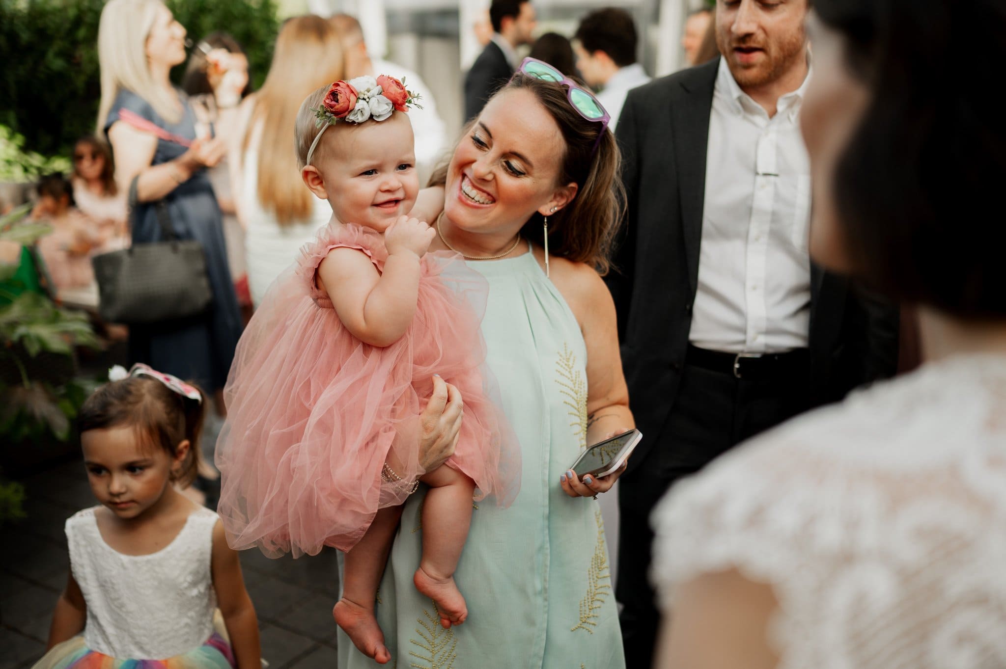 At an outdoor event, a smiling woman holds a baby in a pink dress amid a group of people. The joyful scene captures the happiness and togetherness that are often seen in wedding photos at Blockhouse.