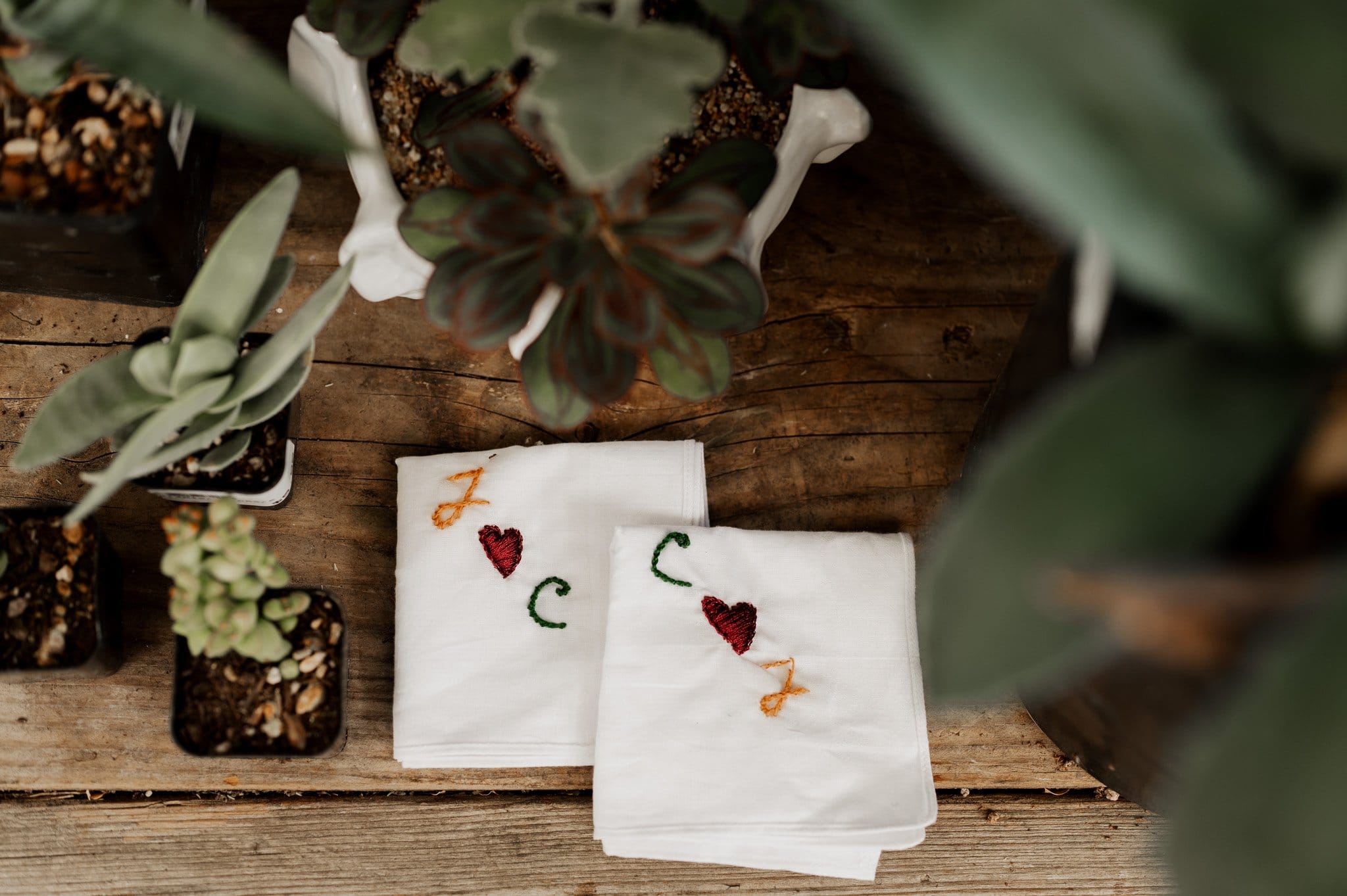 Two embroidered cloths with hearts and initials, surrounded by potted succulents on a wooden surface—perfect touches for wedding photos at Blockhouse.