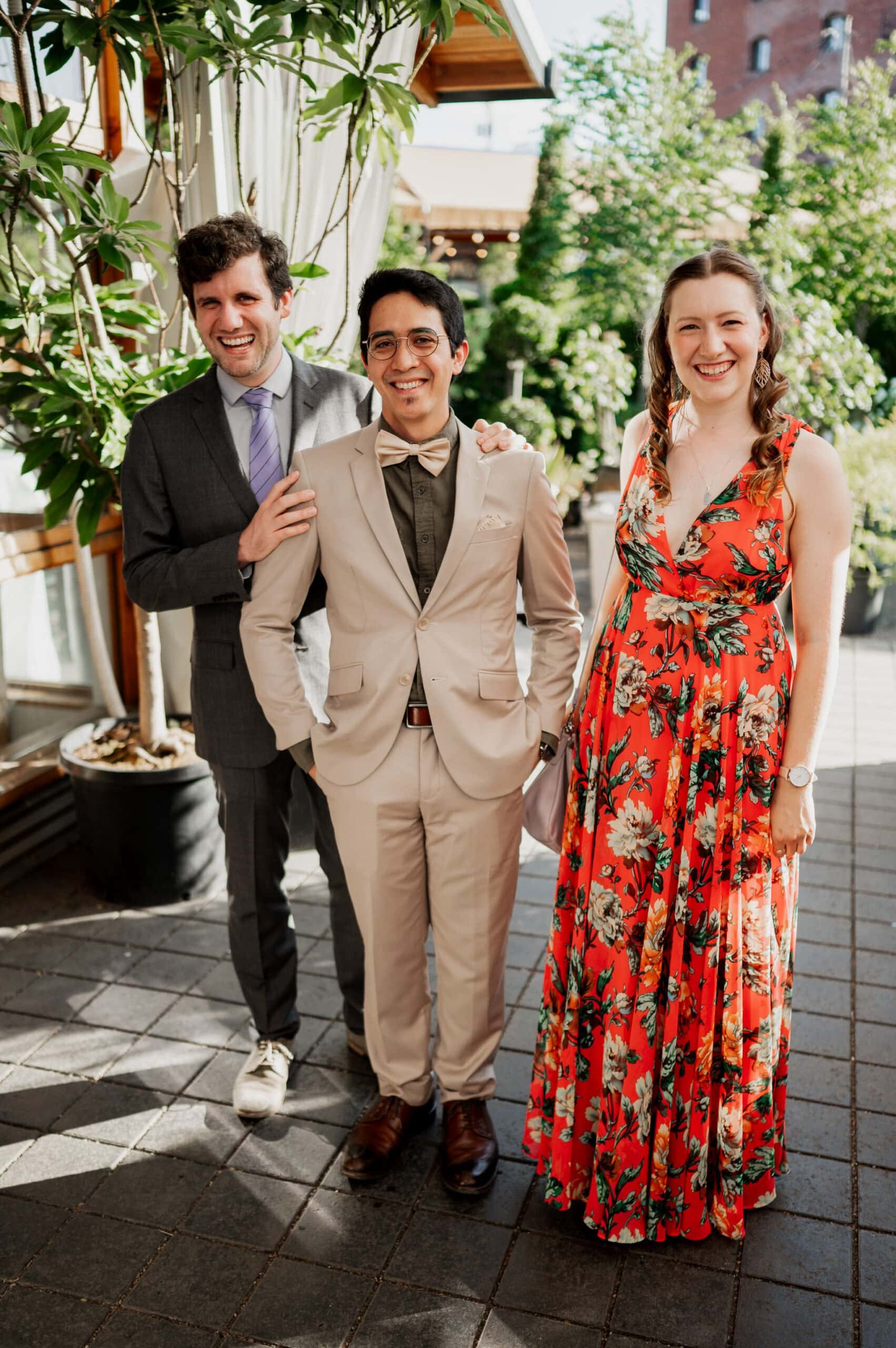 Three people smiling, standing outdoors in a picturesque setting; one in a gray suit, another in a beige suit, and a person in a floral dress. Their joy is perfectly captured in these wedding photos at Blockhouse.