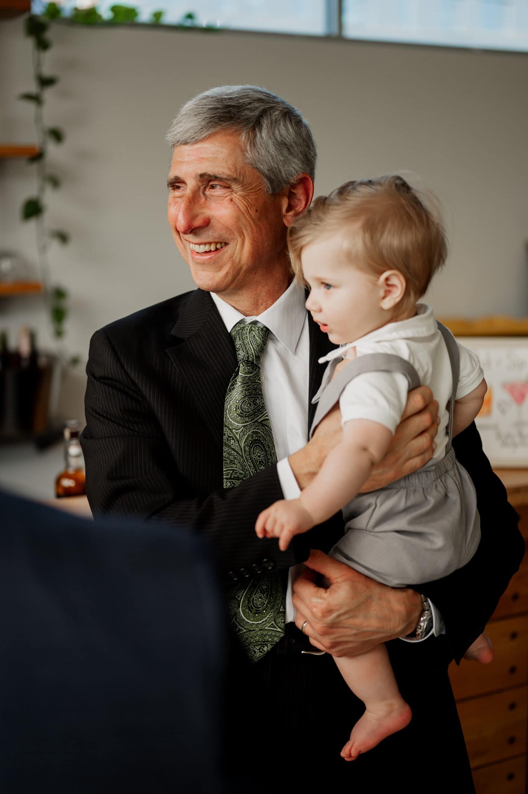 An elderly man in a suit smiles warmly as he holds a toddler in a light outfit, standing amidst shelves and plants. Their joyful connection is as memorable as cherished wedding photos at Blockhouse.