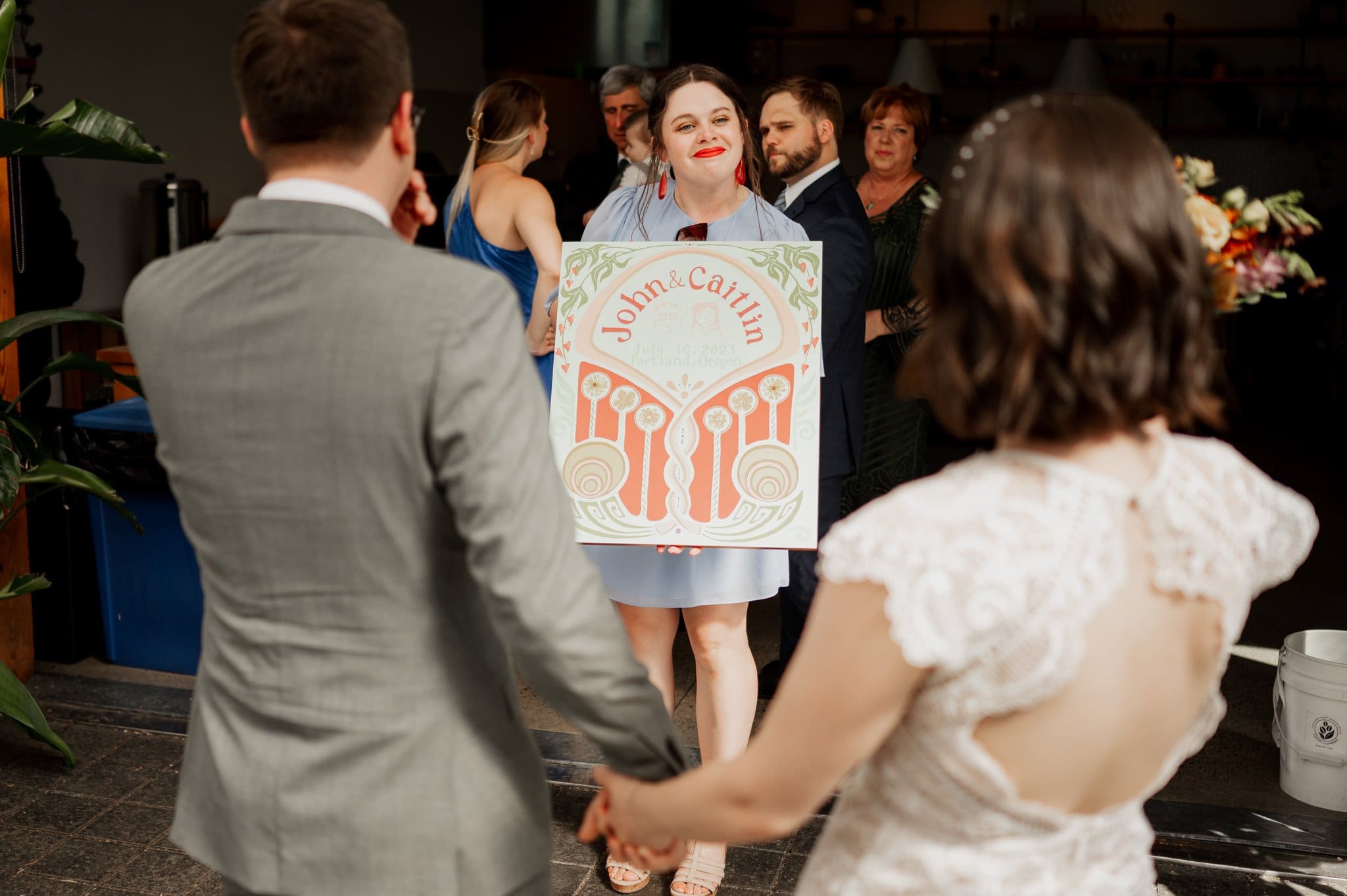 A woman holds a colorful sign, facing a couple holding hands at their wedding event, perhaps capturing wedding photos at Blockhouse.