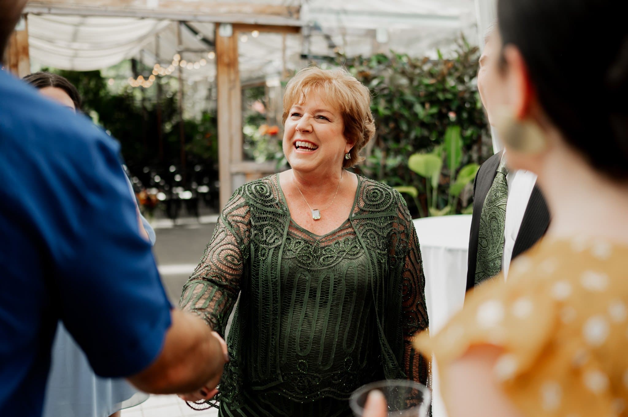 A smiling woman in a green dress greets someone with a handshake at an outdoor gathering, capturing the essence of wedding photos at Blockhouse.