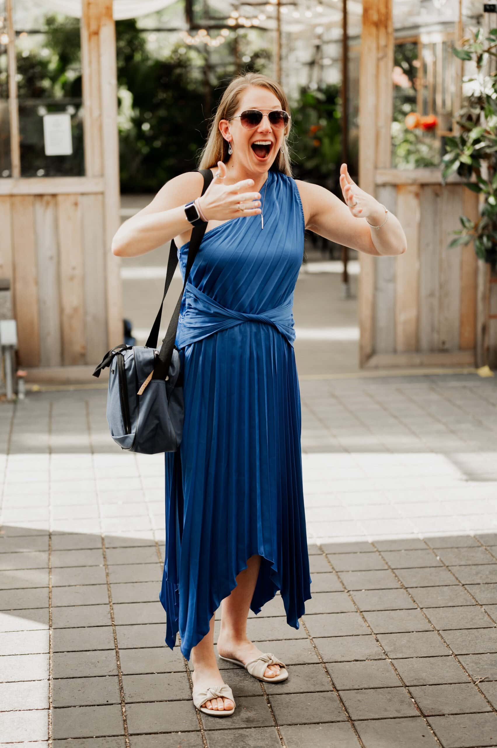 A woman in a blue dress and sandals gestures excitedly outdoors, wearing sunglasses and a shoulder bag, as she poses gracefully for wedding photos at Blockhouse.