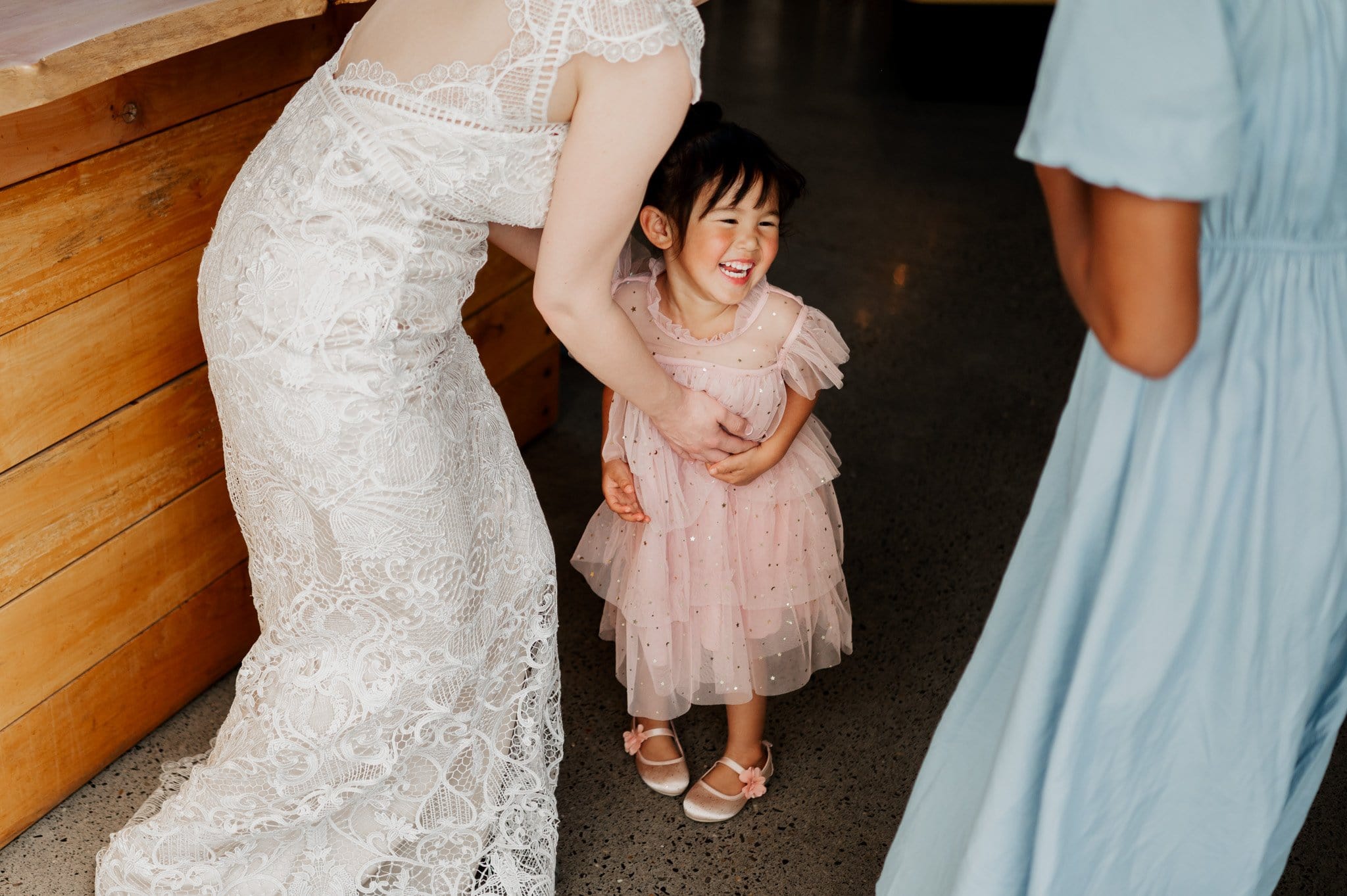 A smiling child in a pink dress is embraced by a woman in a white lace dress, with another in blue nearby. Captured at the picturesque Blockhouse, this moment adds warmth to their wedding photos.