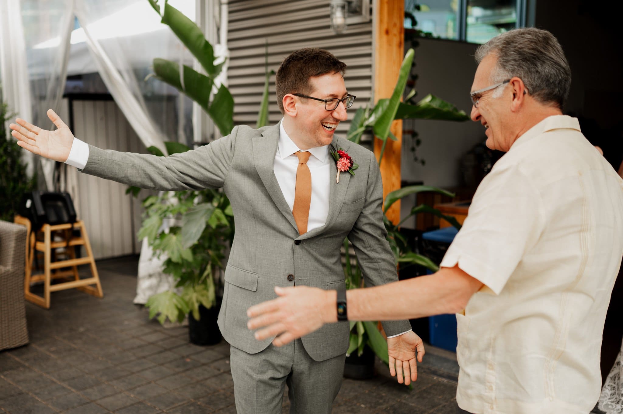 Two men smiling and about to hug, one in a gray suit with a pink boutonniere, the other in a casual shirt. Their joyful connection adds warmth to the wedding photos at Blockhouse.