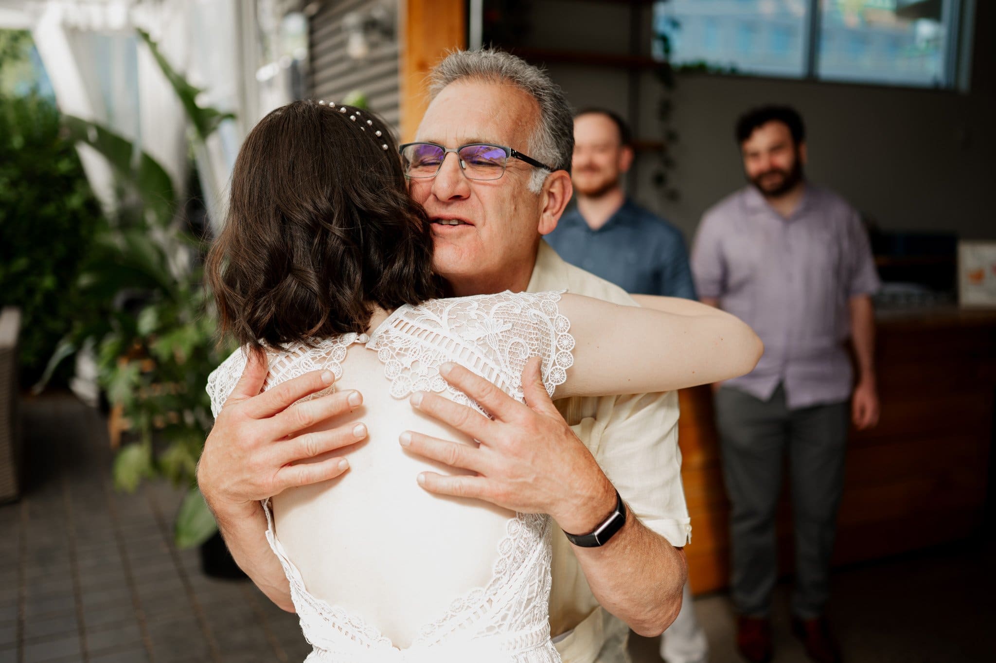 A man joyfully hugs a woman in a white dress at what appears to be a wedding, with two other men smiling in the background. This delightful moment is captured beautifully as part of wedding photos at Blockhouse.