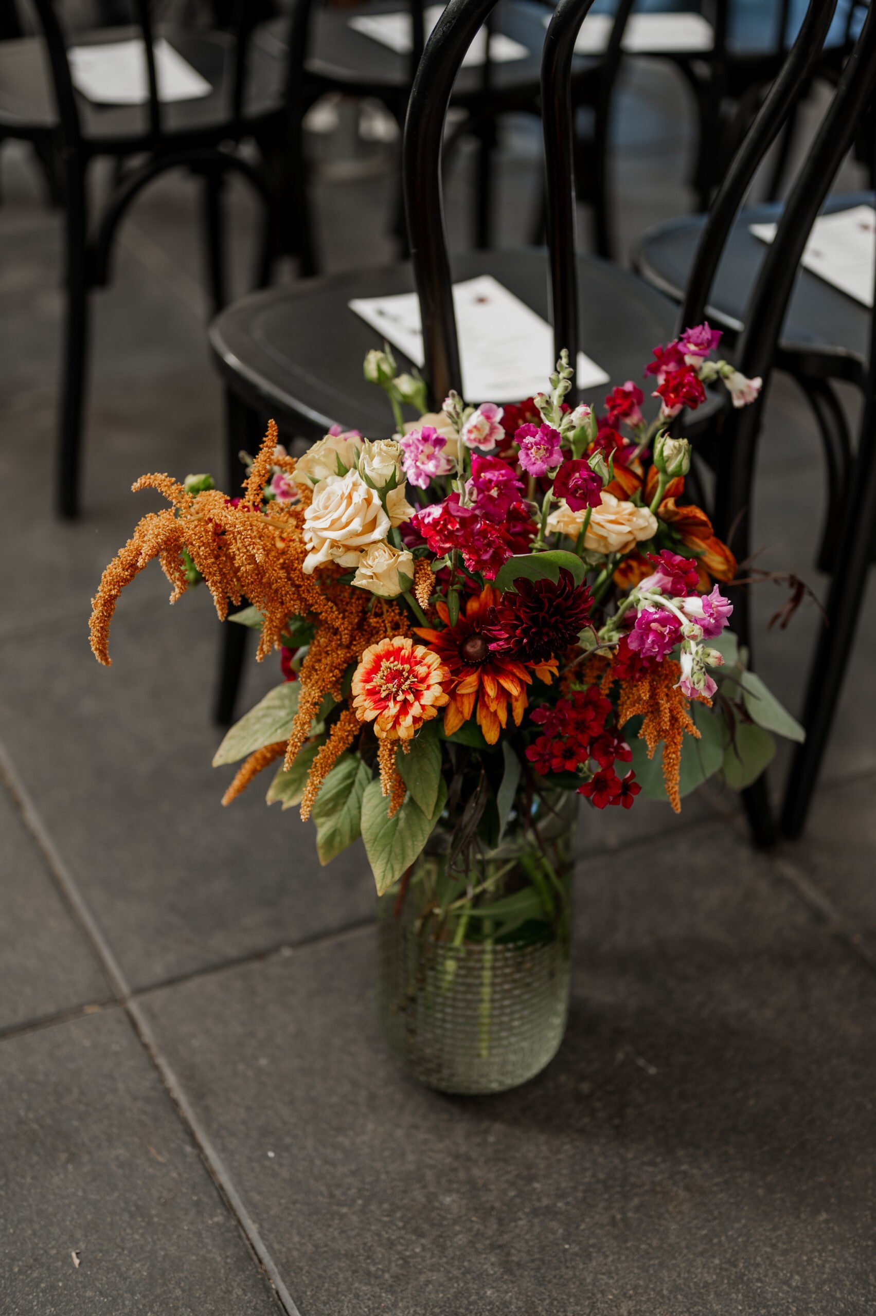 A stunning floral arrangement featuring orange, pink, and cream flowers graces a clear vase on the floor, nestled by black chairs. Perfect for enhancing wedding photos at Blockhouse.