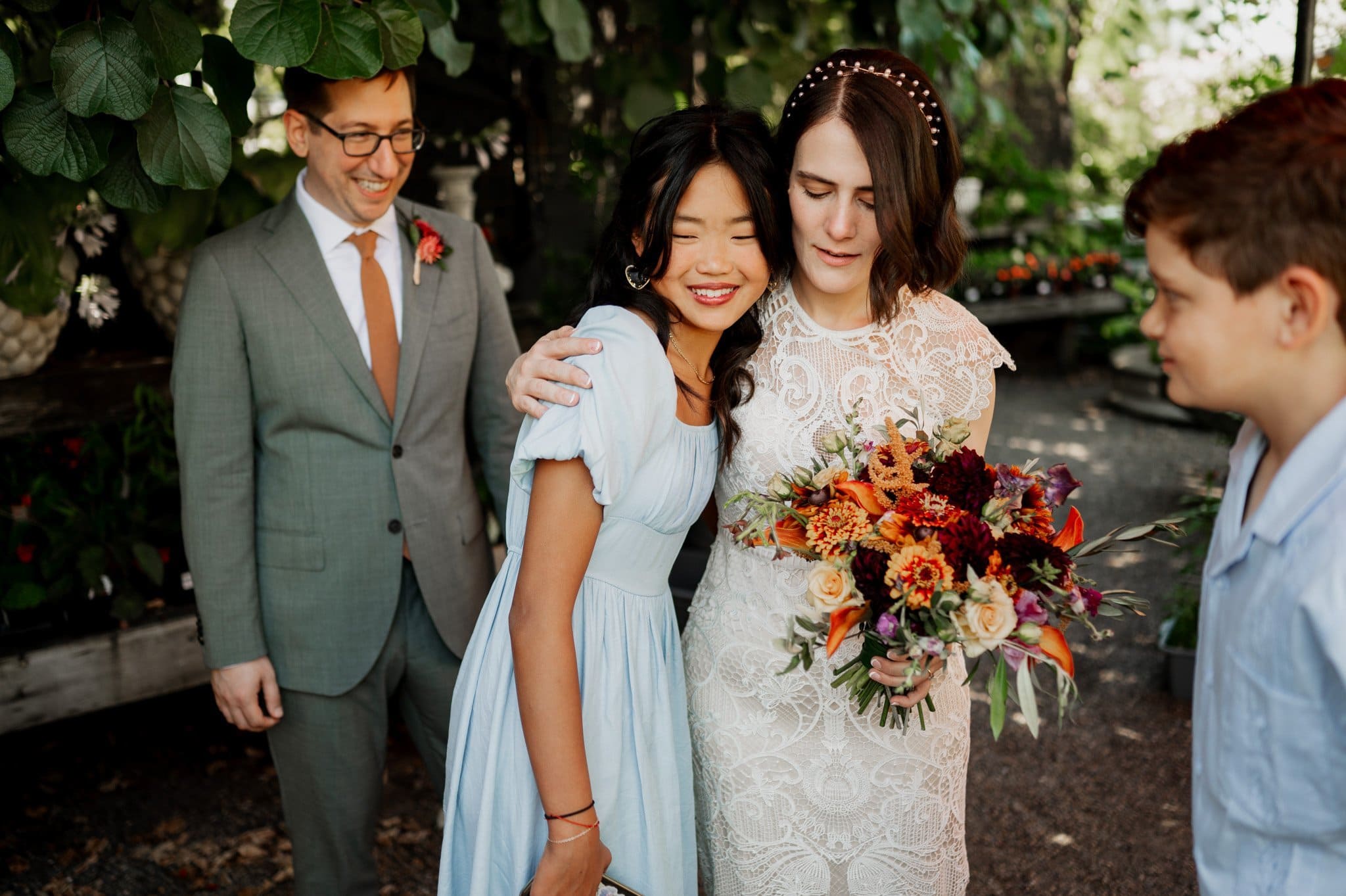 The bride, in her white dress, clutches vibrant flowers as she embraces a woman in light blue. Nearby, two men join them, all smiling warmly under the open sky. Its a perfect moment captured among the wedding photos at Blockhouse.