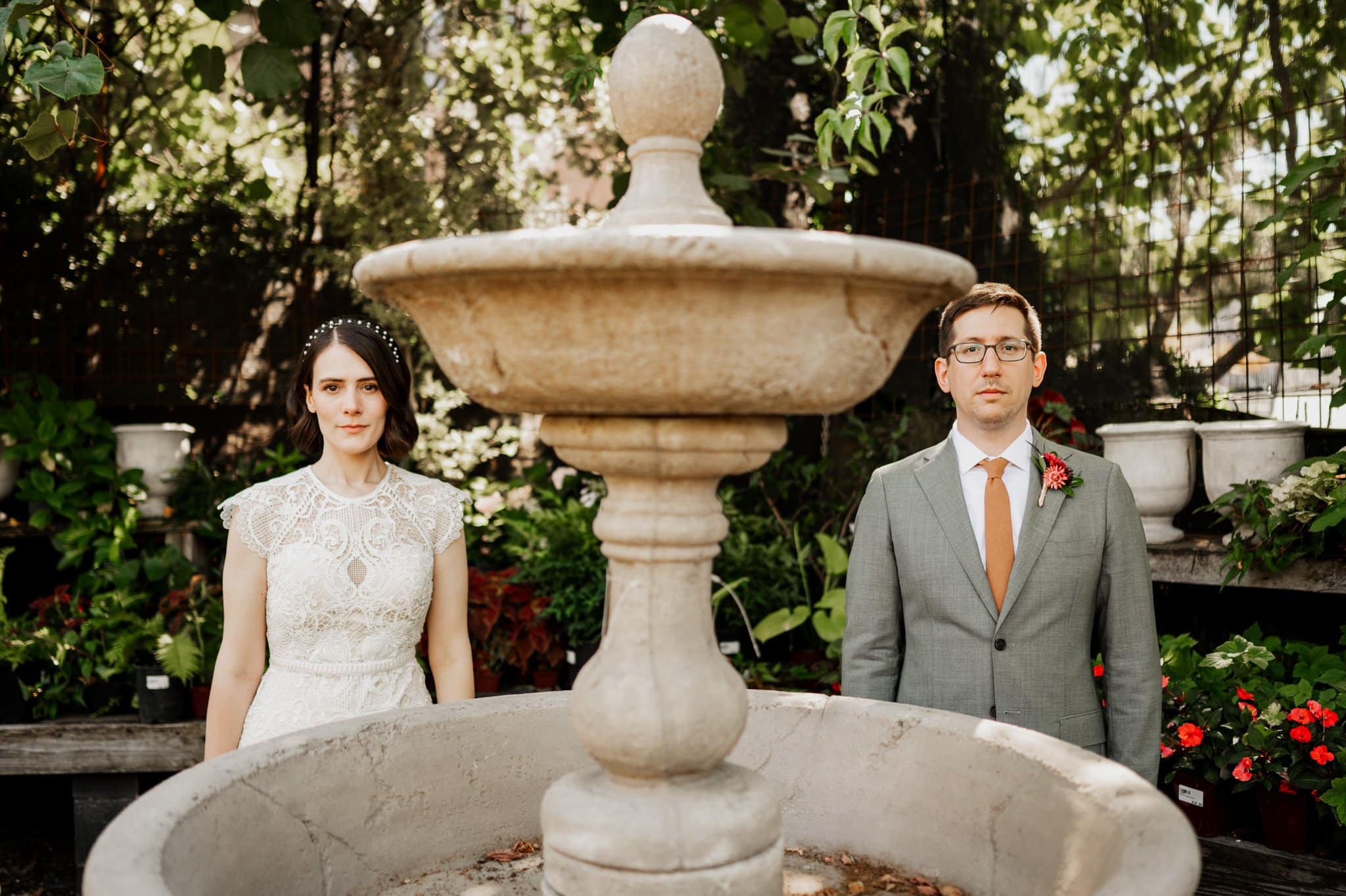 The bride and groom pose elegantly behind a stone fountain, surrounded by the lush greenery of a garden during their wedding photos at Blockhouse.