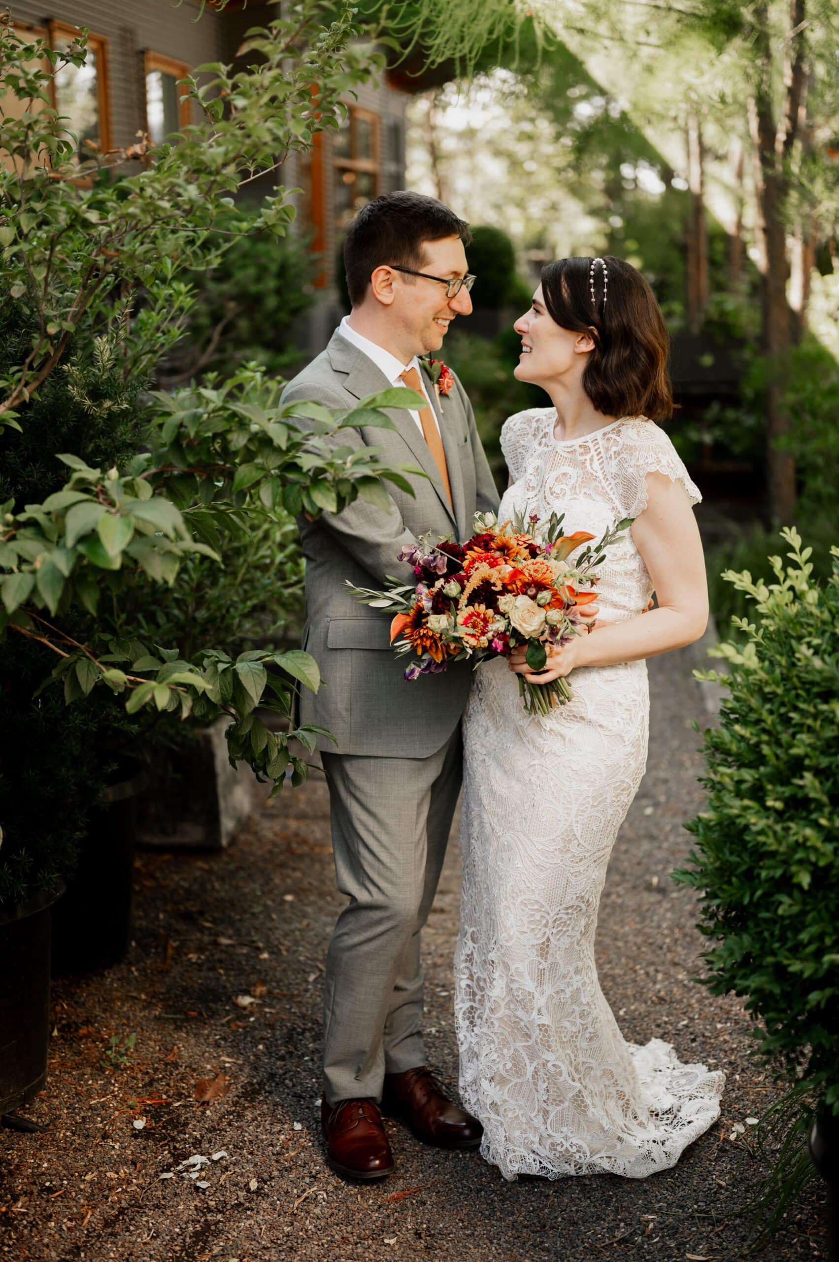 The bride and groom smile at each other, surrounded by lush greenery, with the bride holding a colorful bouquet. Their joy is beautifully captured in these wedding photos at Blockhouse.