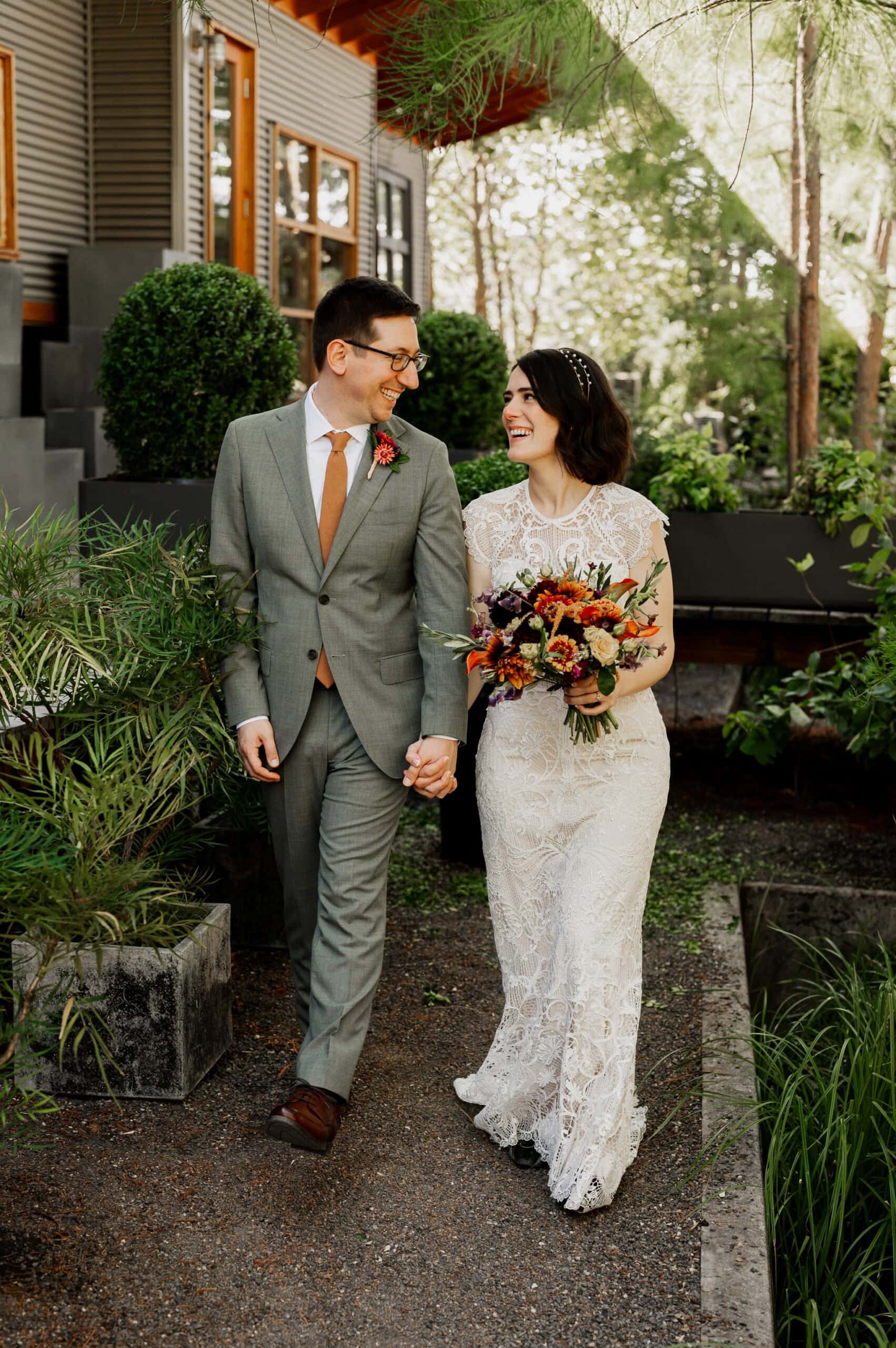 The couple, smiling and holding hands, strolls outside during their wedding photos at Blockhouse. The brides lace gown complements her colorful bouquet beautifully.