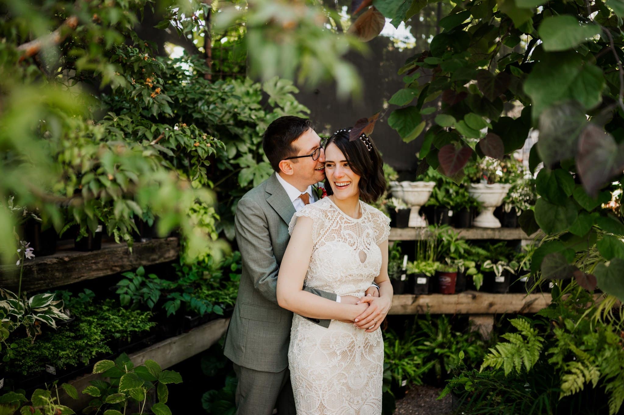 A couple embraces and smiles in a lush garden at Blockhouse, with the woman in a white dress and the man in a gray suit.
