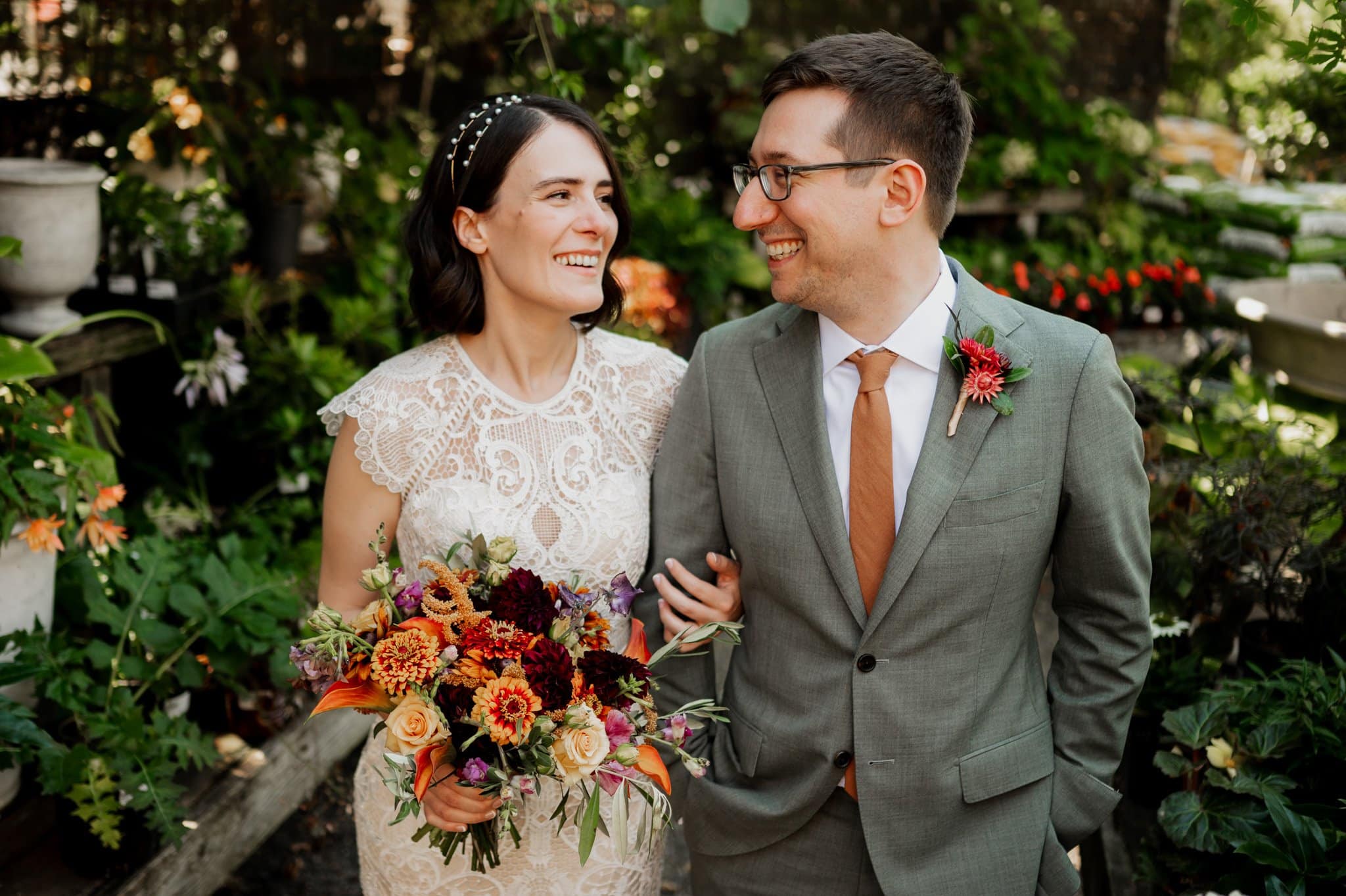 The bride in a lace dress and groom in a gray suit share a smile, holding a colorful bouquet amidst lush greenery. A perfect moment captured during their wedding photos at Blockhouse.