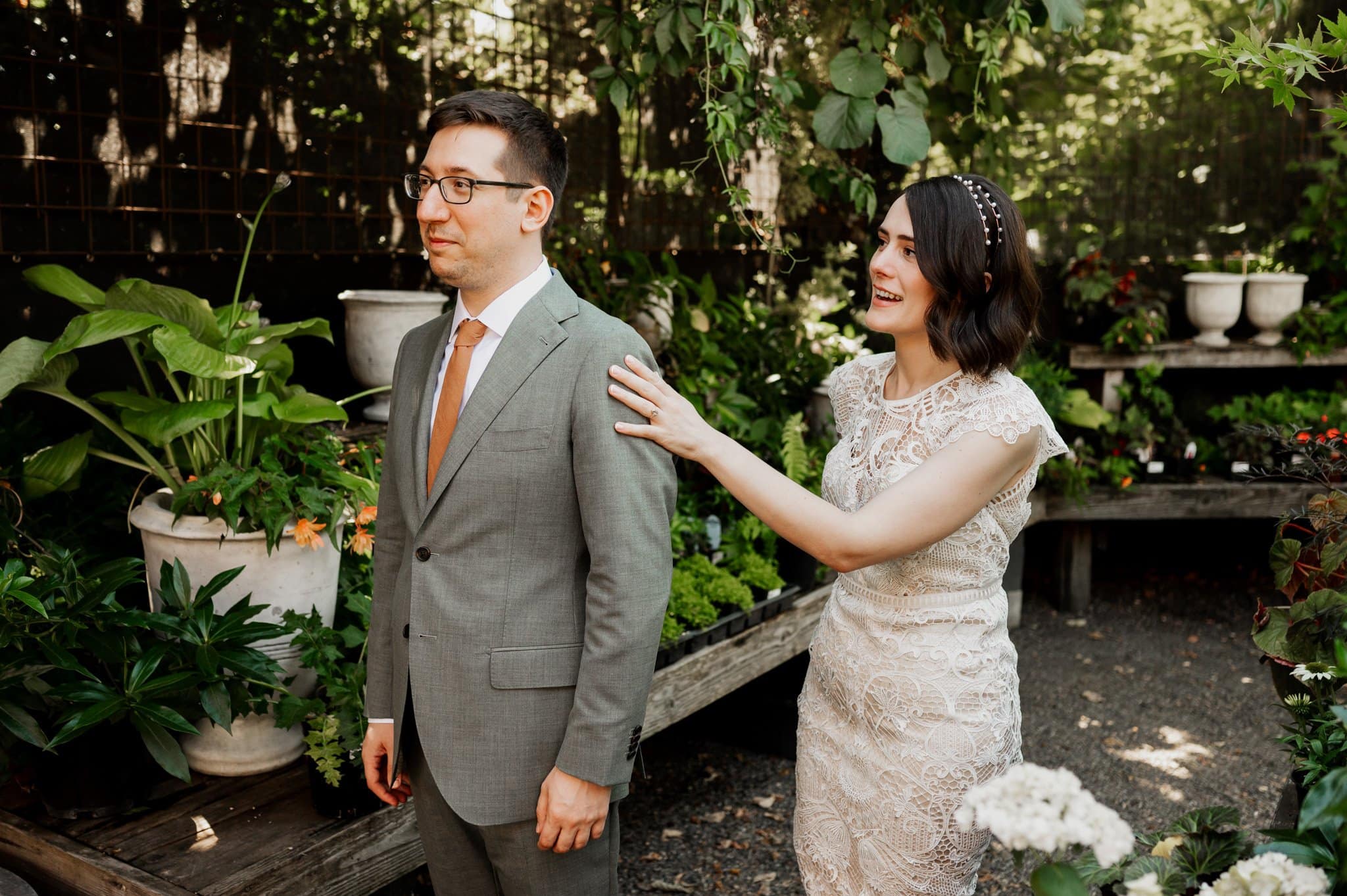 Bride in a lace dress surprises groom in a gray suit amid lush garden plants, perfect for wedding photos at Blockhouse.
