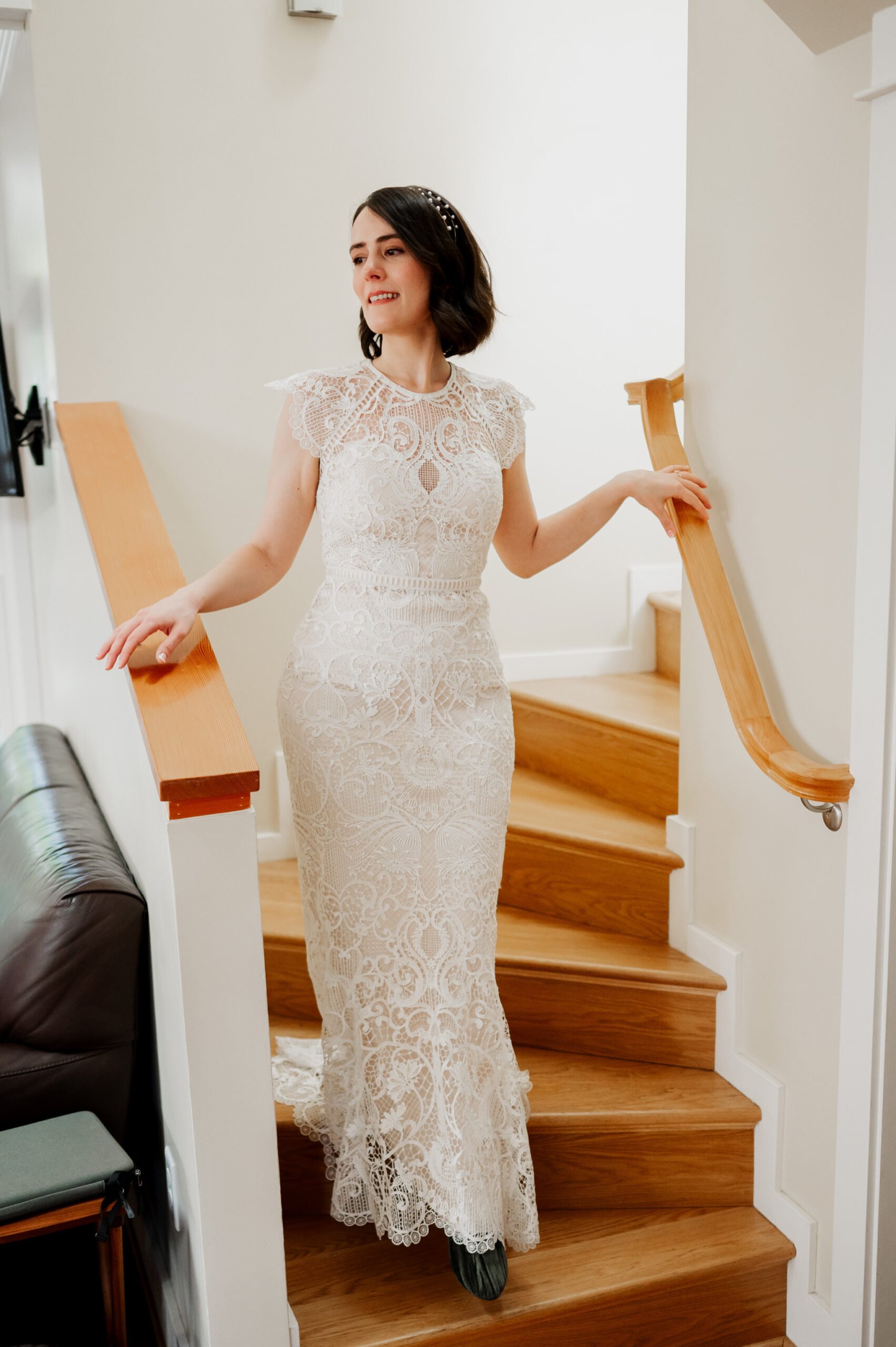 A woman in a lace wedding dress stands on wooden stairs in a room with white walls, getting ready at an Airbnb for her wedding at Blockhouse.