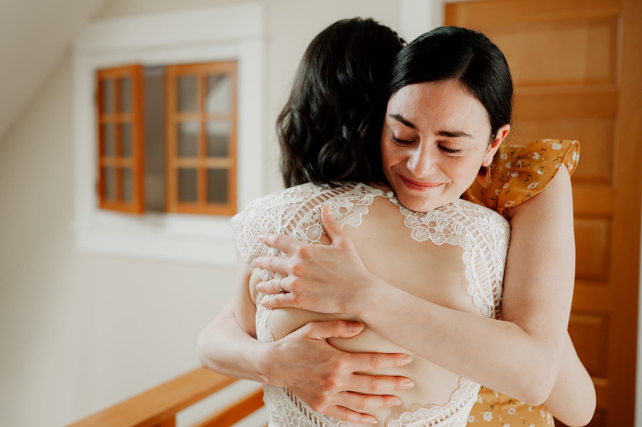 Two women hugging warmly indoors, one in a white lace dress and the other in mustard yellow, as they get ready at an Airbnb for a wedding at Blockhouse.