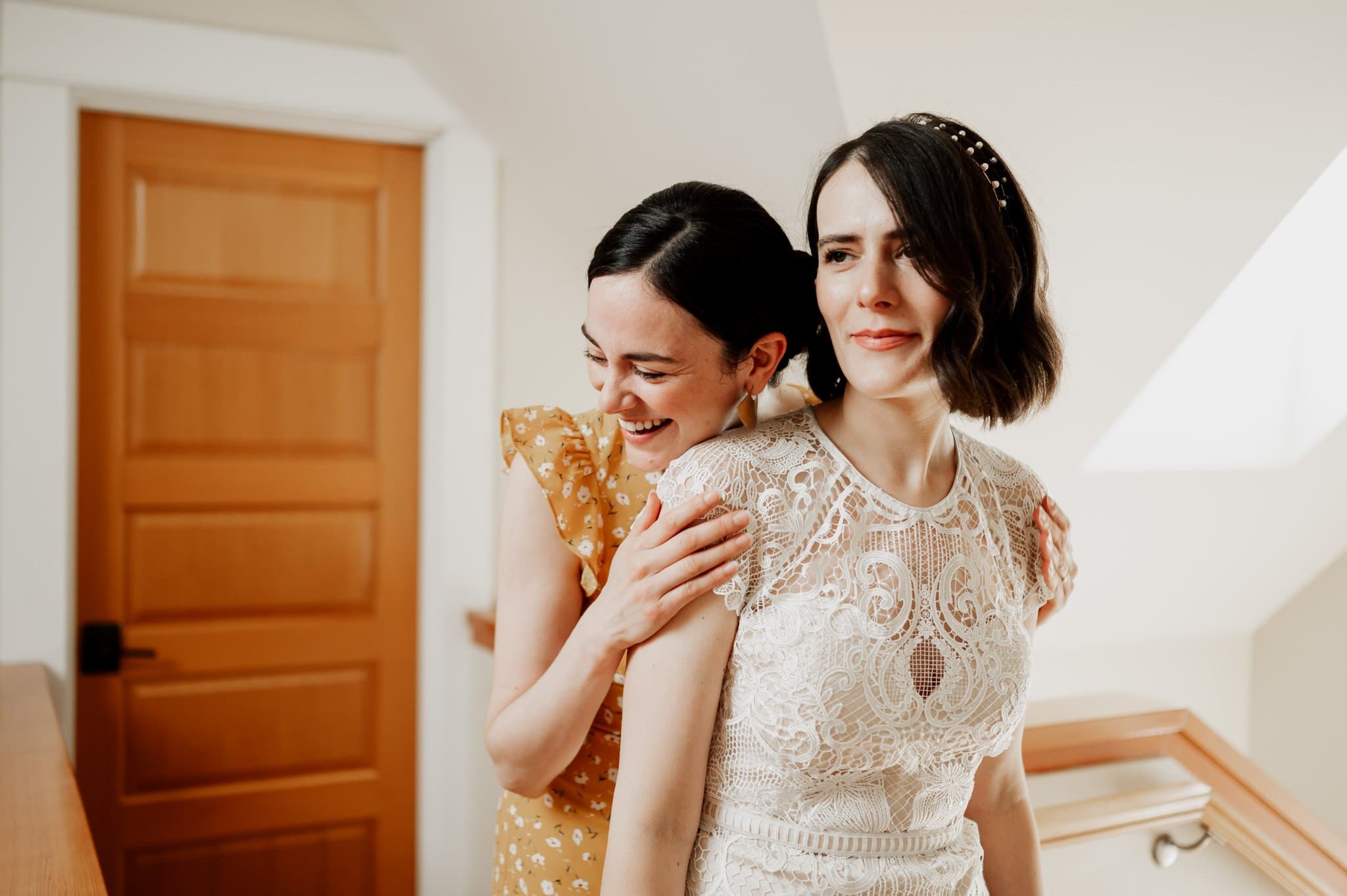 Two women smiling; one in a lace dress, the other in a yellow dress, standing near a wooden door, getting ready at an Airbnb for a wedding at Blockhouse.