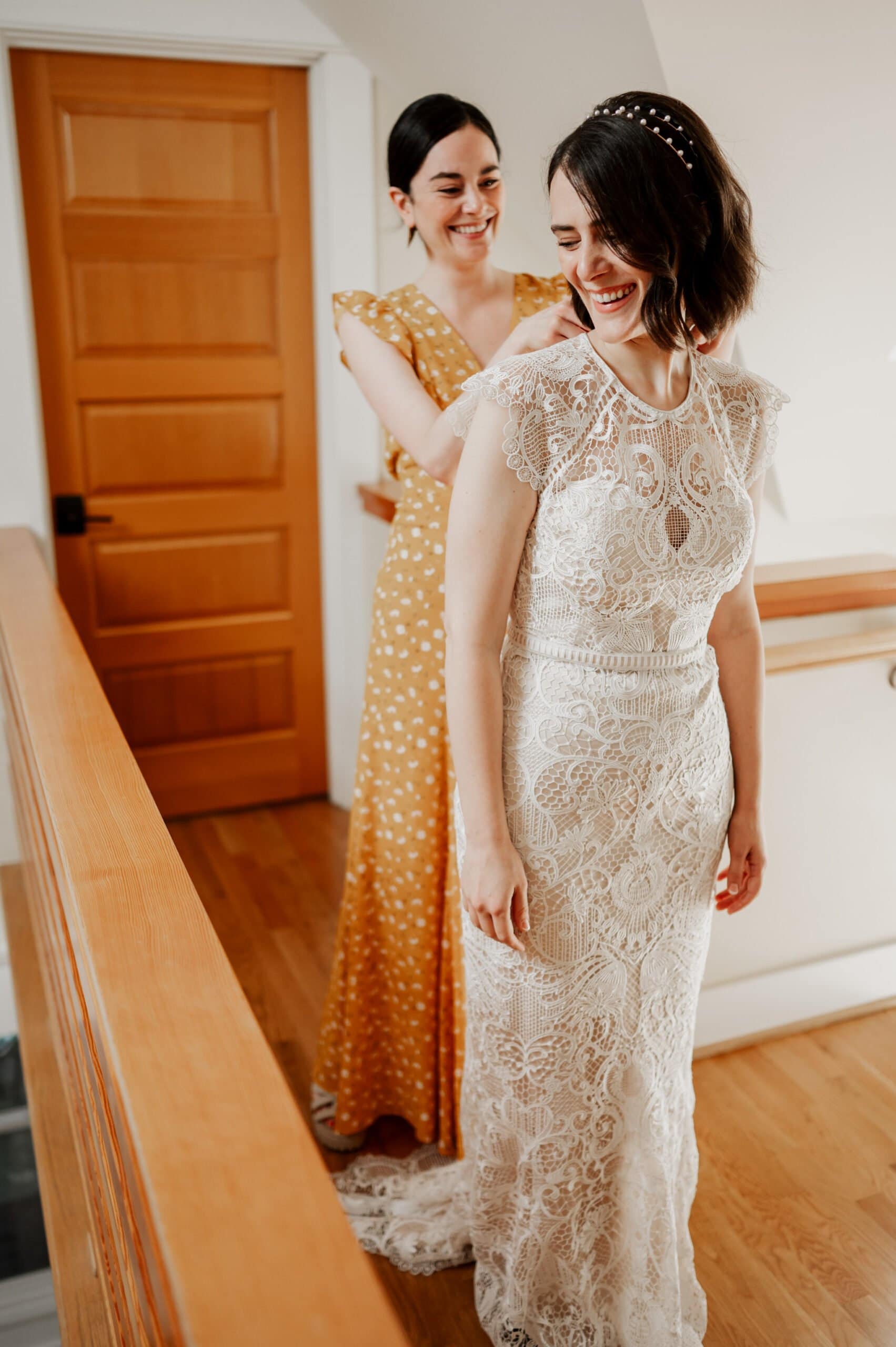A bride in a lace gown smiles as a woman in a yellow dress assists her indoors, getting ready for the wedding at Blockhouse.