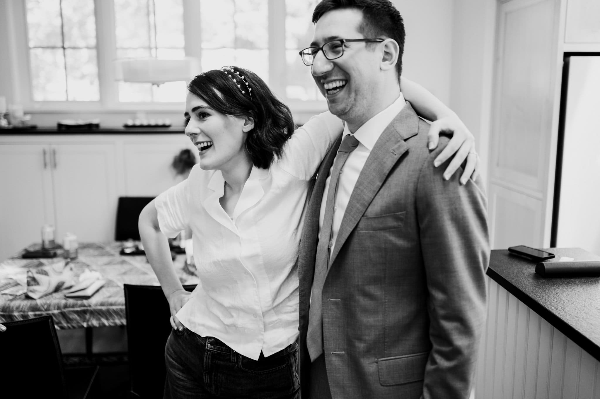 A smiling couple stands together, with the woman embracing the man as they look towards the left, getting ready at an Airbnb for a wedding at Blockhouse.