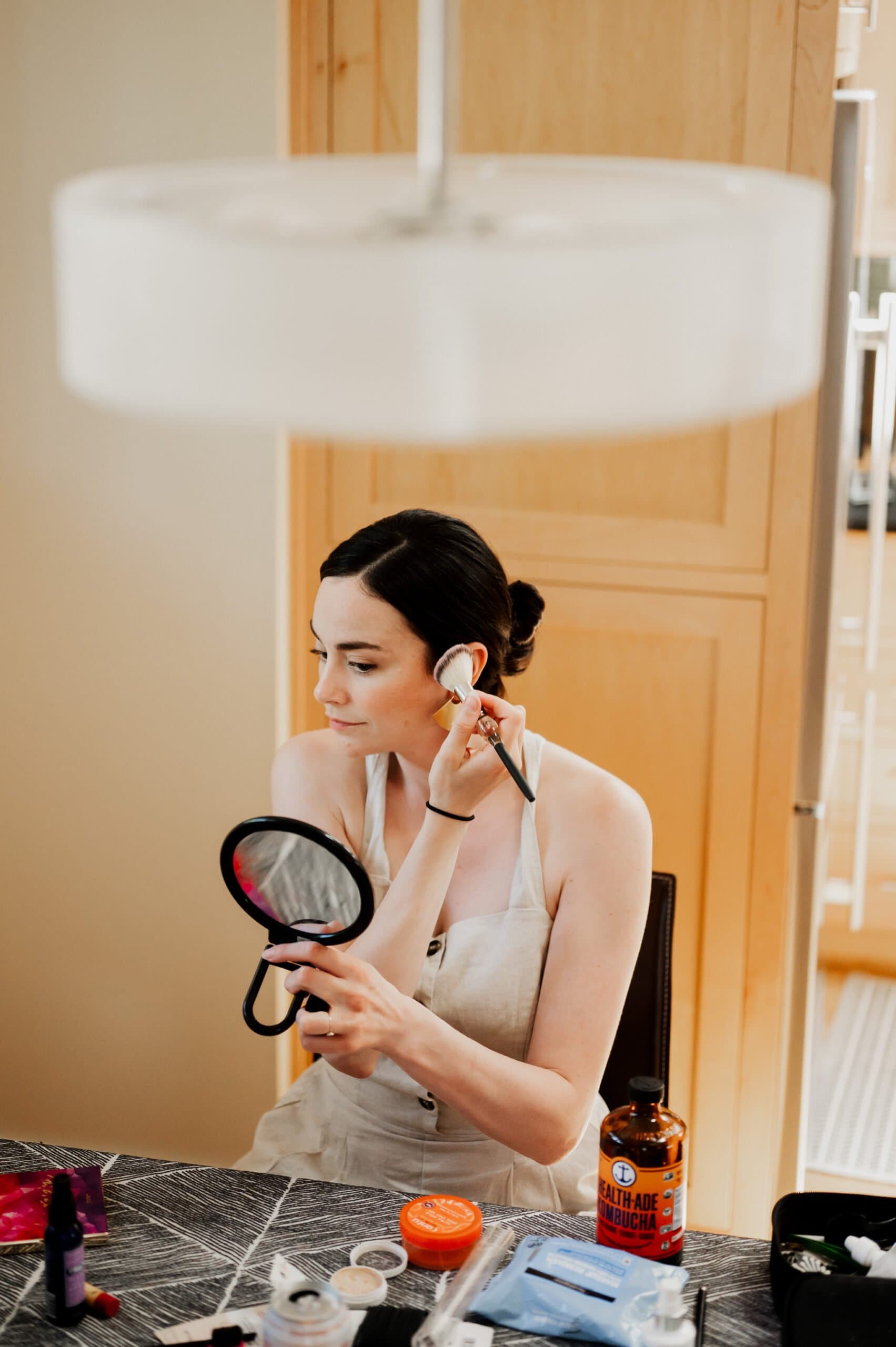 The woman is getting ready at an Airbnb for a wedding at Blockhouse, applying makeup with a brush and checking her reflection in the mirror. She sits at a table surrounded by beauty products, focused on perfecting her look.