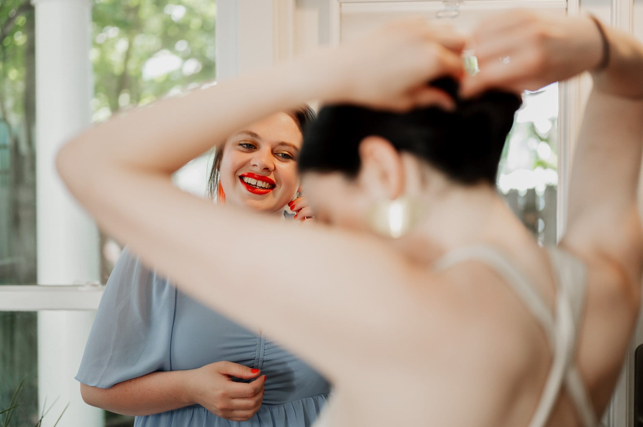 A woman in a blue dress smiles while another adjusts her hair, both getting ready at an Airbnb for a wedding at Blockhouse.