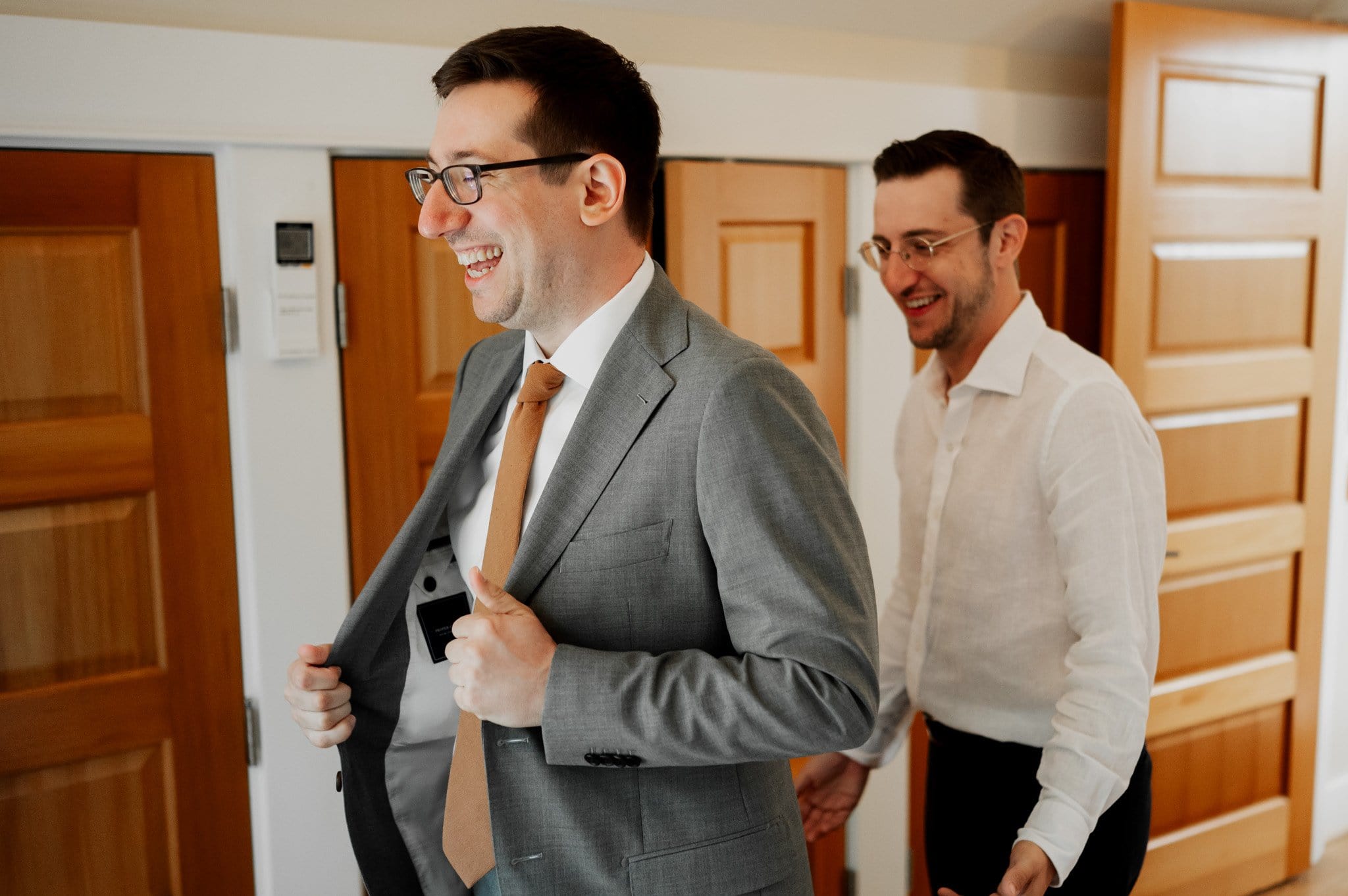 Two men smiling, one in a gray suit adjusting his jacket, the other in a white shirt. Theyre getting ready at an Airbnb for a wedding at Blockhouse, with wooden doors in the background.