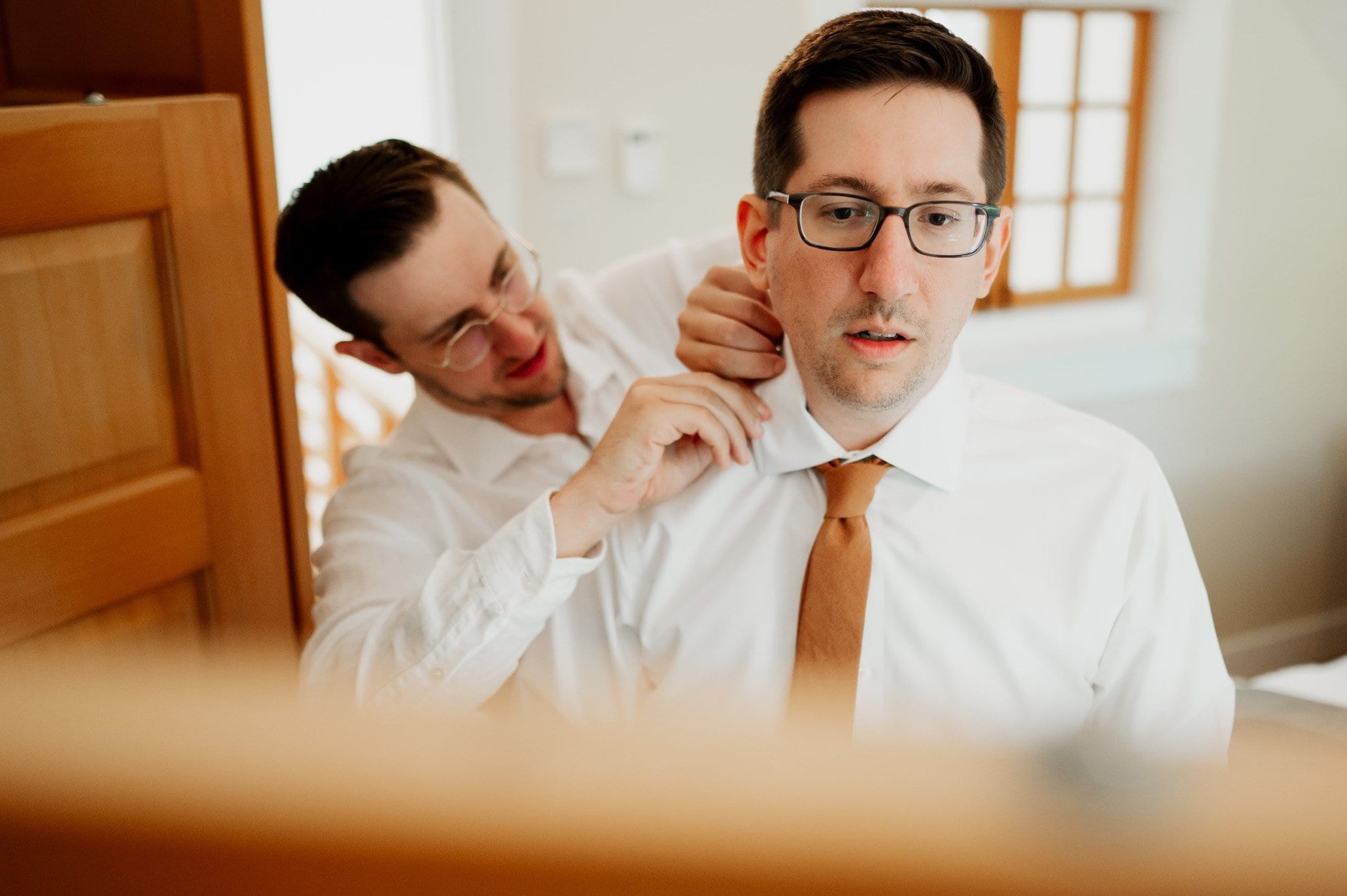 Two men in white shirts are getting ready at an Airbnb for a wedding at Blockhouse; one helps the other adjust his tie in a warmly lit room.
