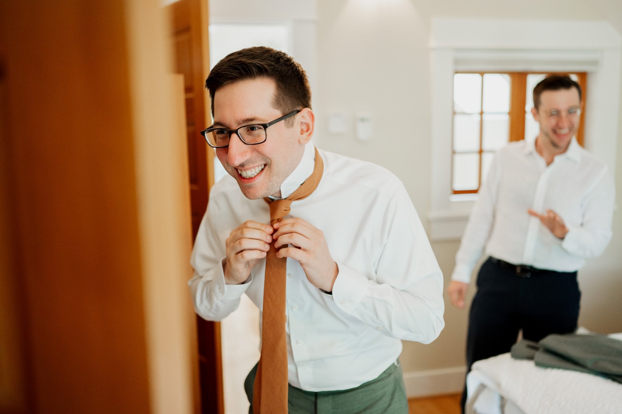 A man in glasses smiles while adjusting his tie, getting ready at an Airbnb for a wedding at Blockhouse; another man in the background shares his cheerful demeanor.