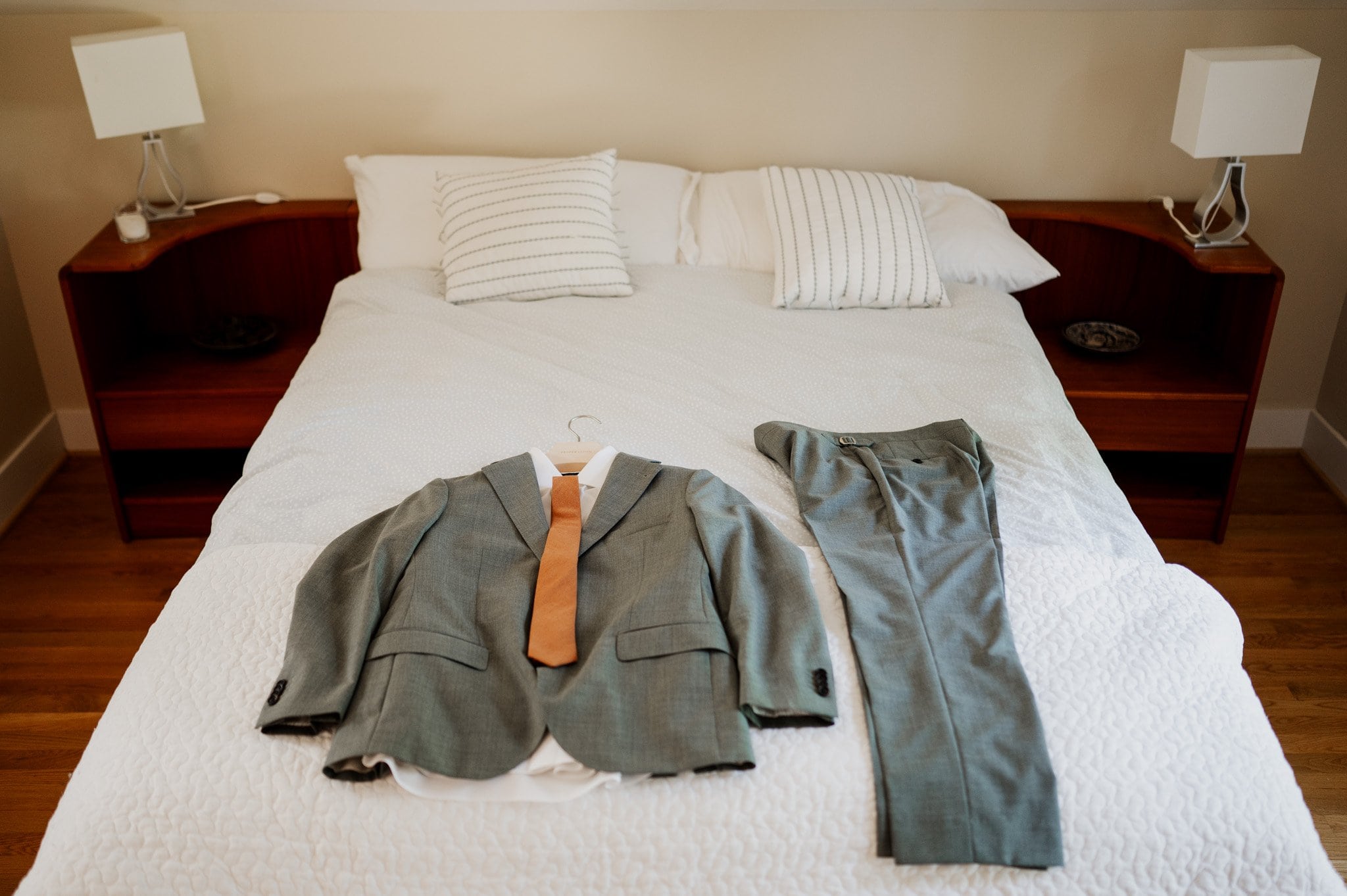 A gray suit, white shirt, and brown tie are laid out on a neatly made bed at an Airbnb near Blockhouse, next to two pillows and bedside tables.