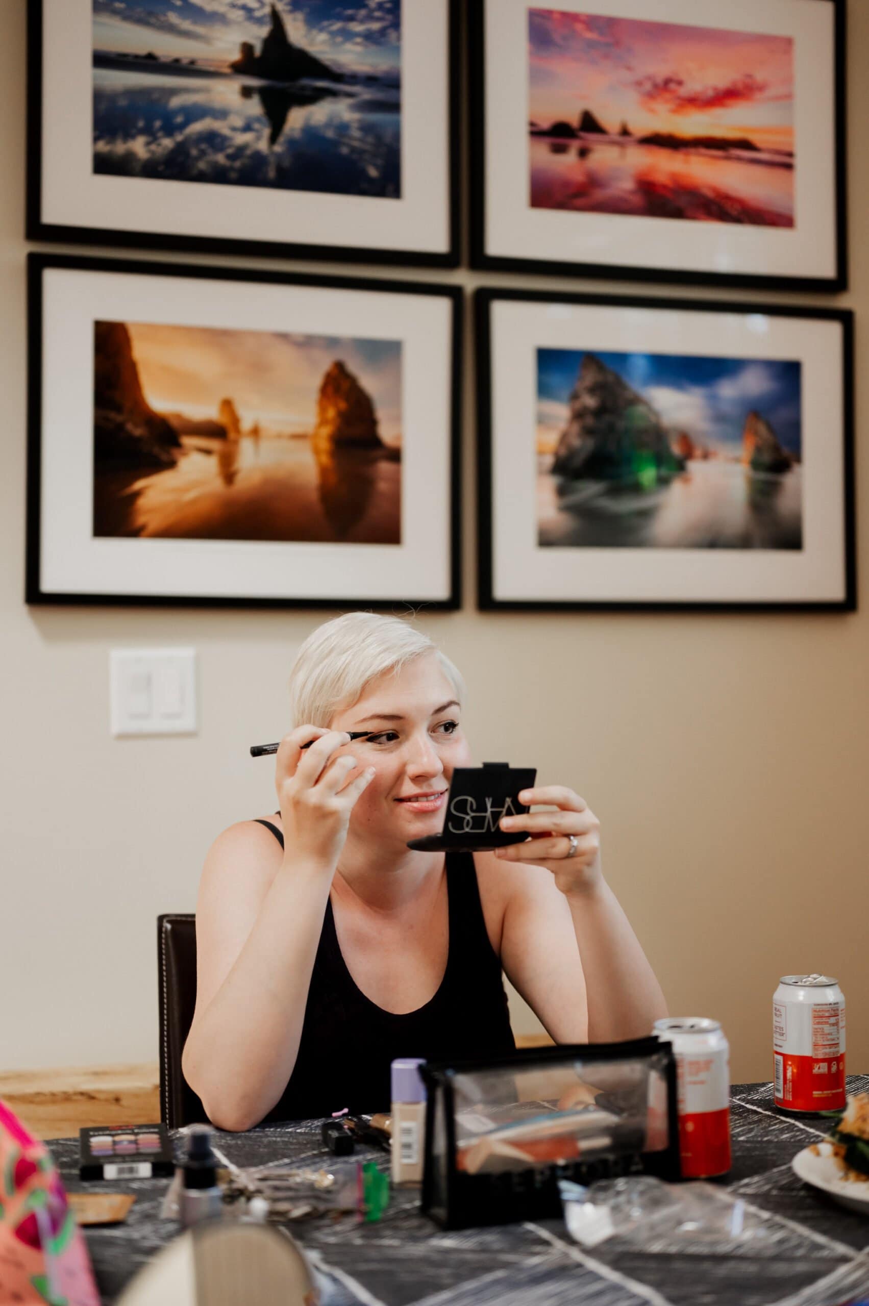 A person is applying makeup at a table, getting ready at an Airbnb for a wedding at Blockhouse, with framed landscape photos on the wall in the background.