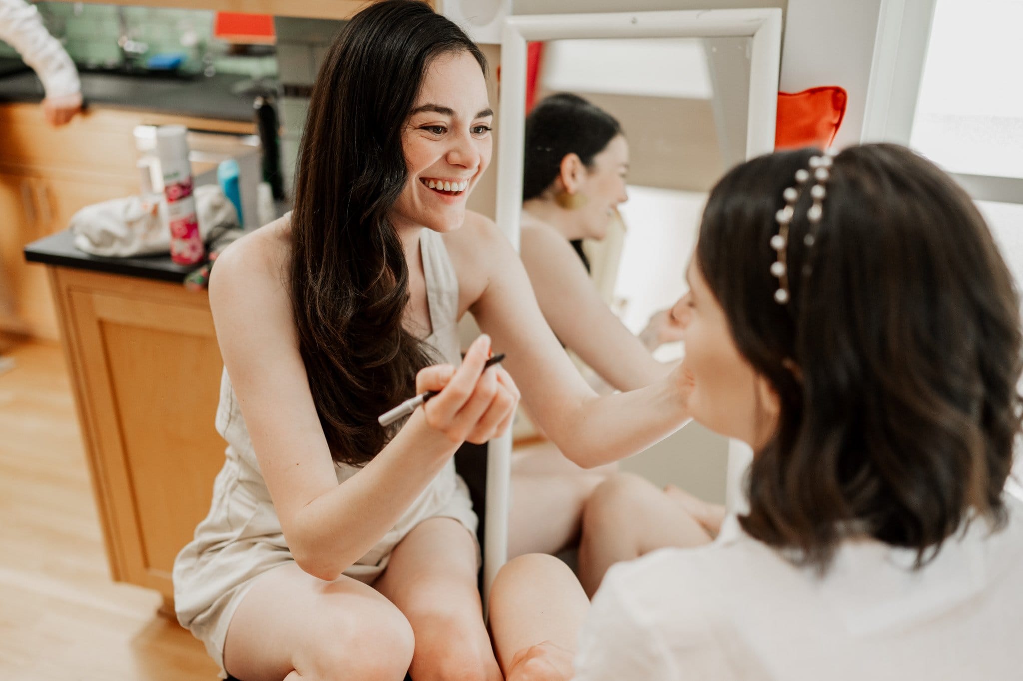 A woman skillfully applies makeup to another woman seated in a chair as they get ready at an Airbnb for a wedding at Blockhouse.