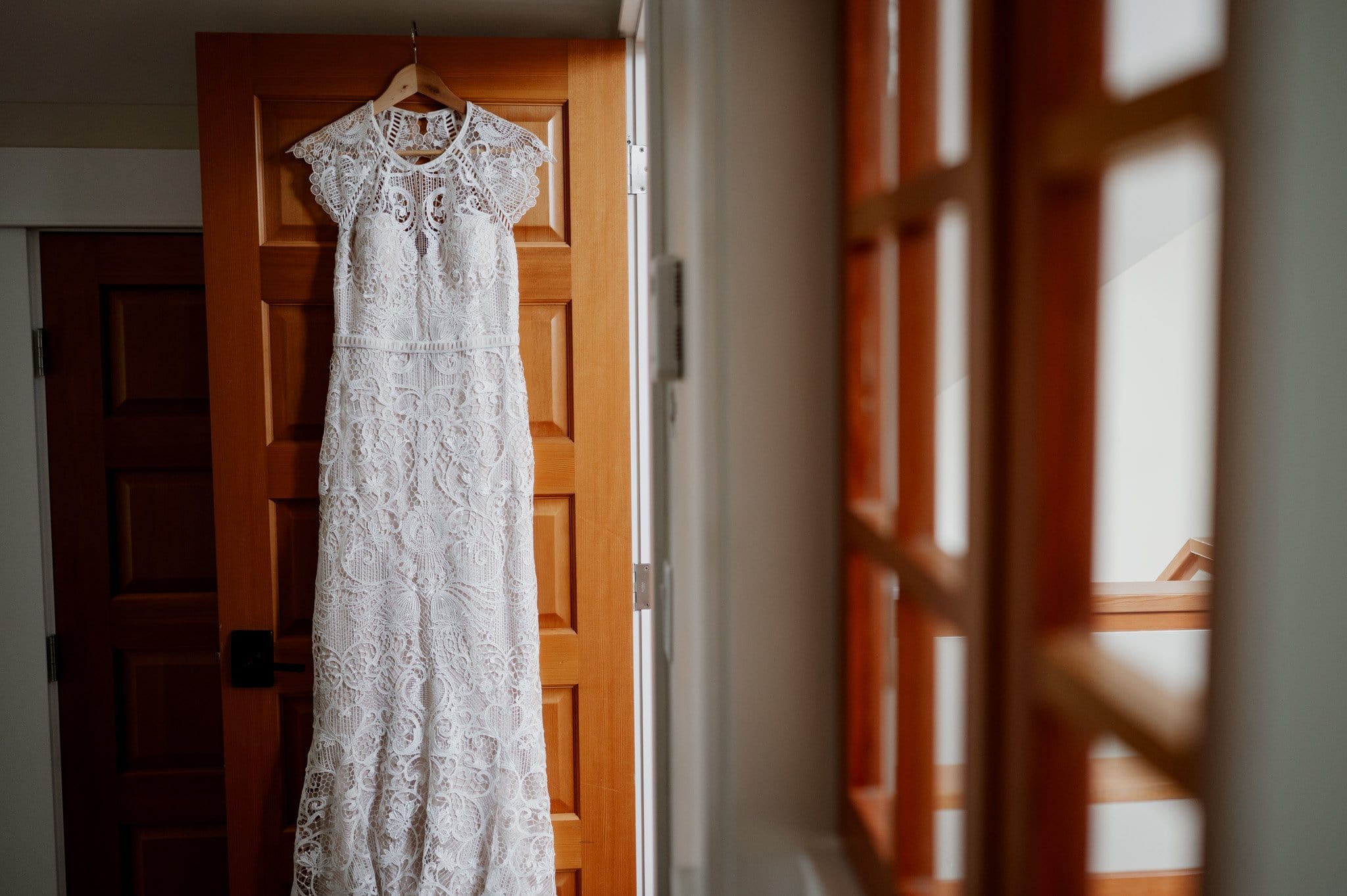 The white lace wedding dress hangs gracefully on a wooden door, softly illuminated by natural light streaming through the adjacent window panes, as the bride is getting ready at an Airbnb for her wedding at Blockhouse.
