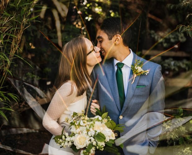 A couple in wedding attire stand closely among greenery, with the bride holding a bouquet and the groom in a blue suit accented by a green tie, capturing timeless Blockhouse wedding photos.