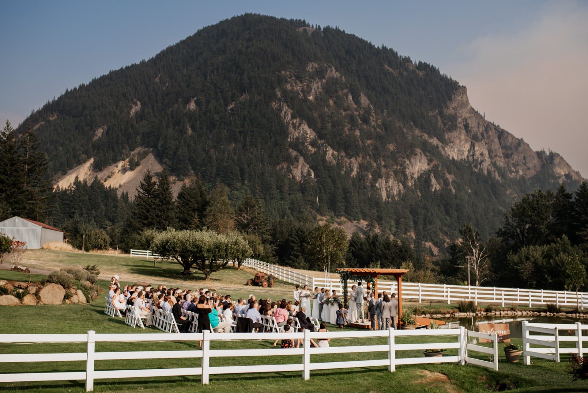 An outdoor wedding ceremony takes place at Wind Mountain Ranch with a mountain backdrop. Guests are seated within a charming white fence that outlines the intimate space.