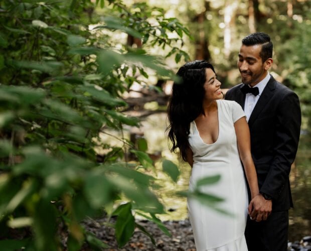 A couple in elegant formal attire smiles warmly at each other while standing in a wooded creek area at Leach Botanical Garden.