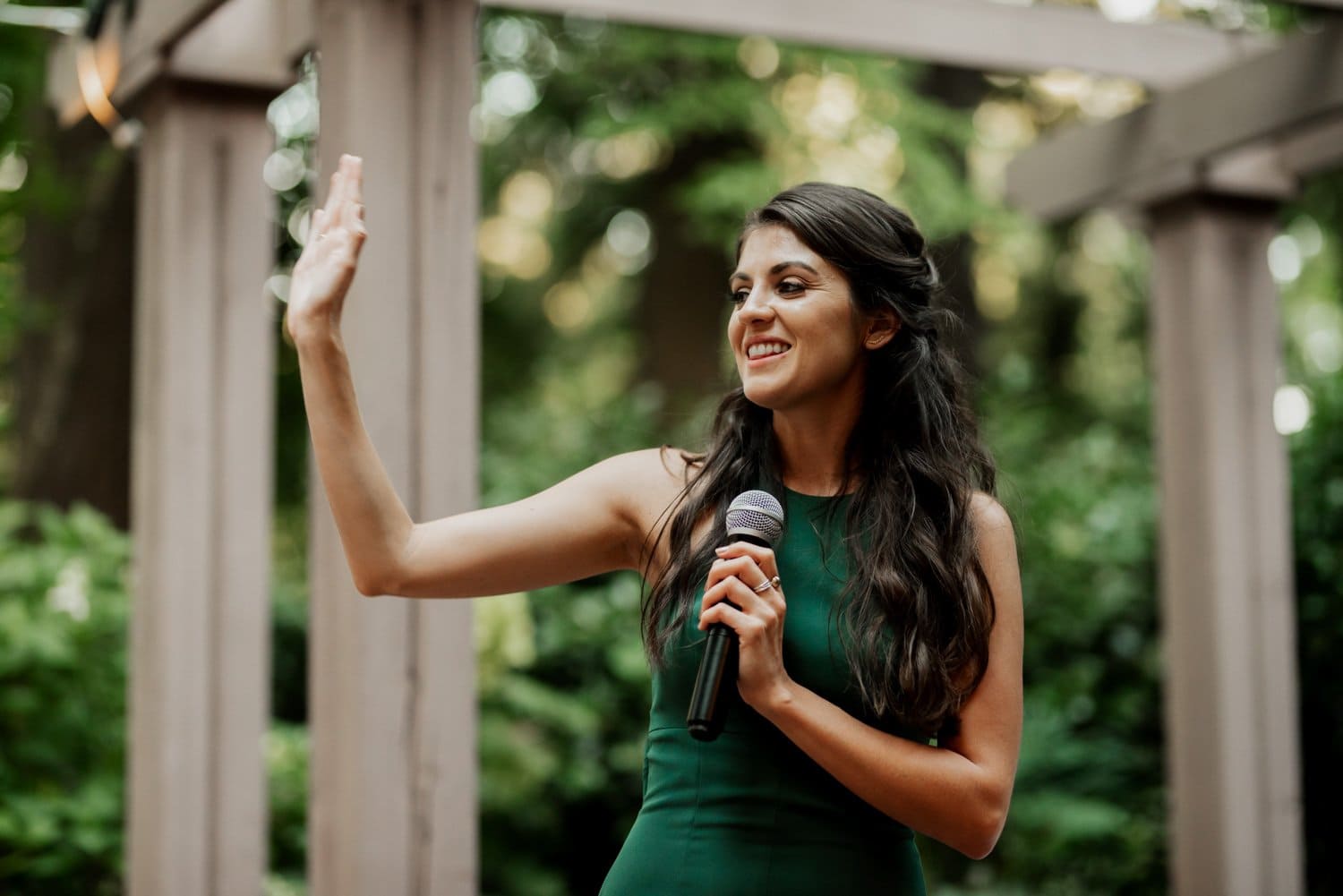 A woman in a green dress waves and holds a microphone, surrounded by the greenery of Leach Botanical Garden.