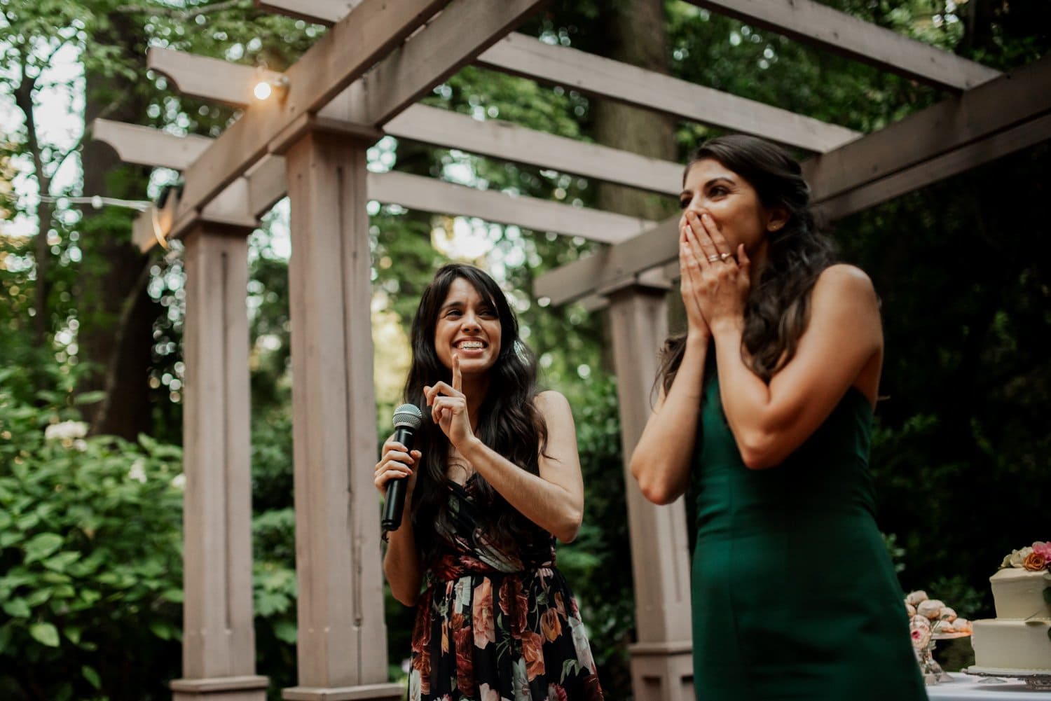 Two women celebrating under a pergola at Leach Botanical Garden, with one holding a microphone and the other smiling and covering her mouth.