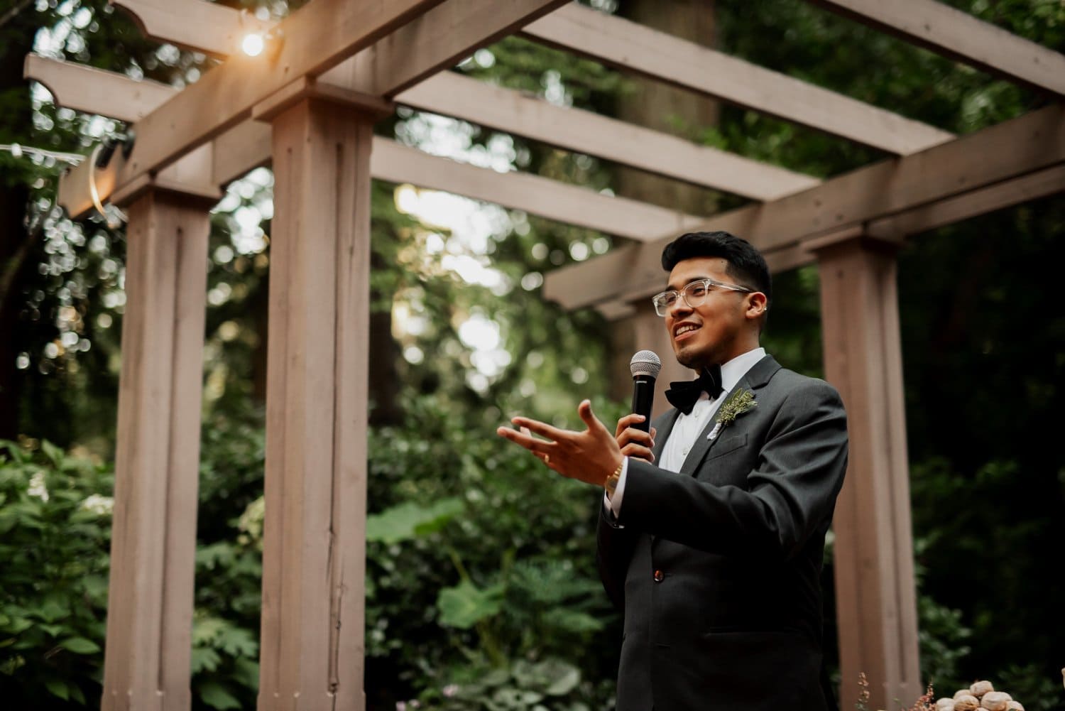 A man in a suit gives a heartfelt speech outdoors under a wooden pergola, surrounded by the lush greenery of Leach Botanical Garden during a wedding celebration.