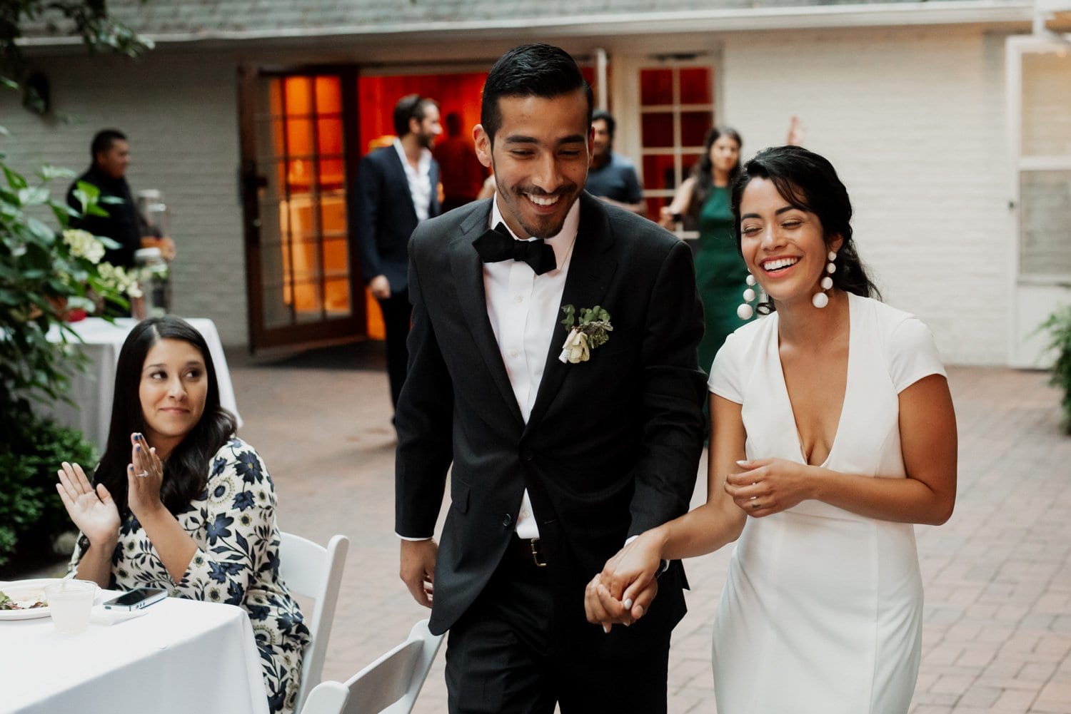A couple in formal attire stands smiling and holding hands at Leach Botanical Garden, surrounded by happy guests.
