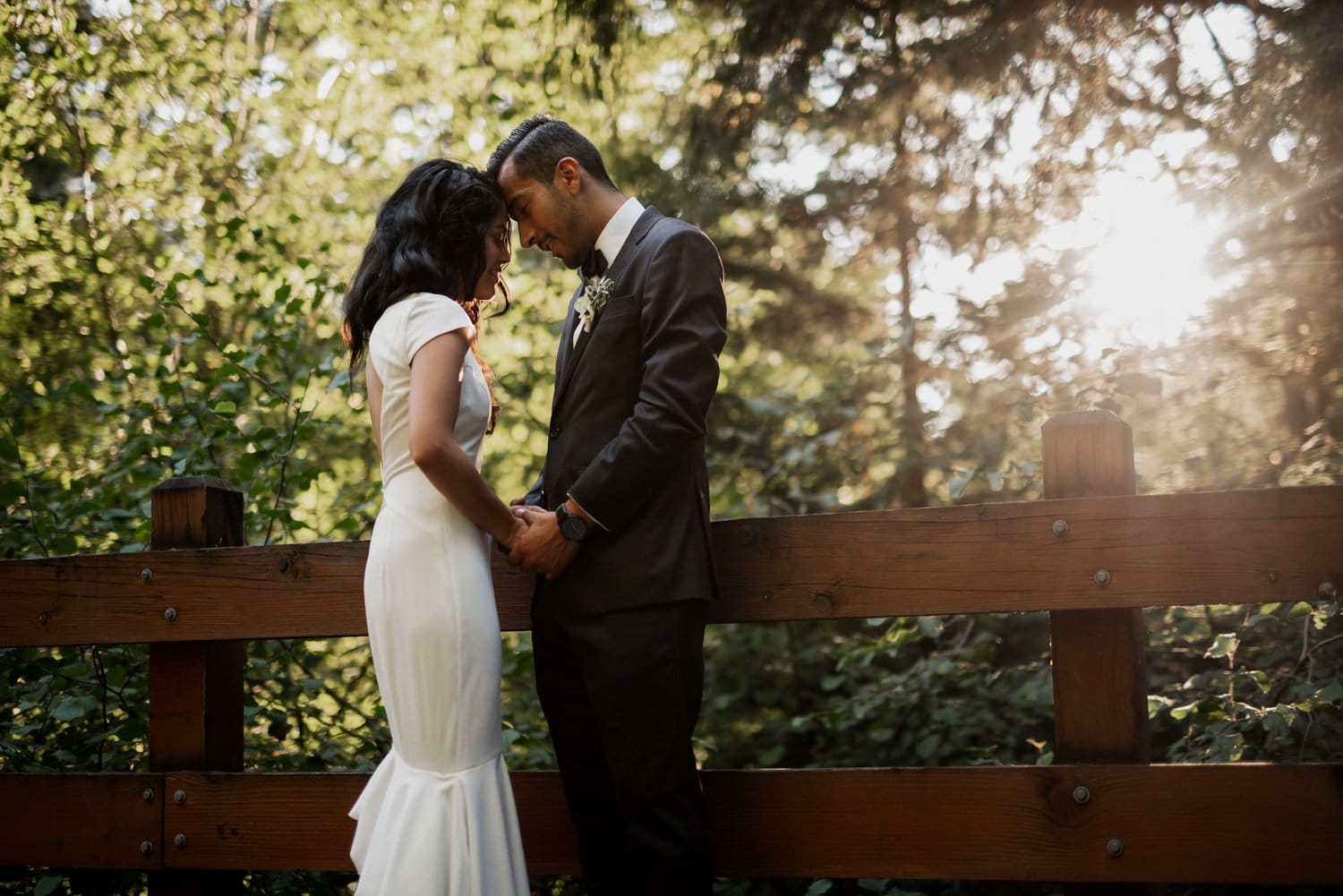 A couple stands closely together on a wooden bridge at Leach Botanical Garden, with sunlight streaming through the trees surrounding them.