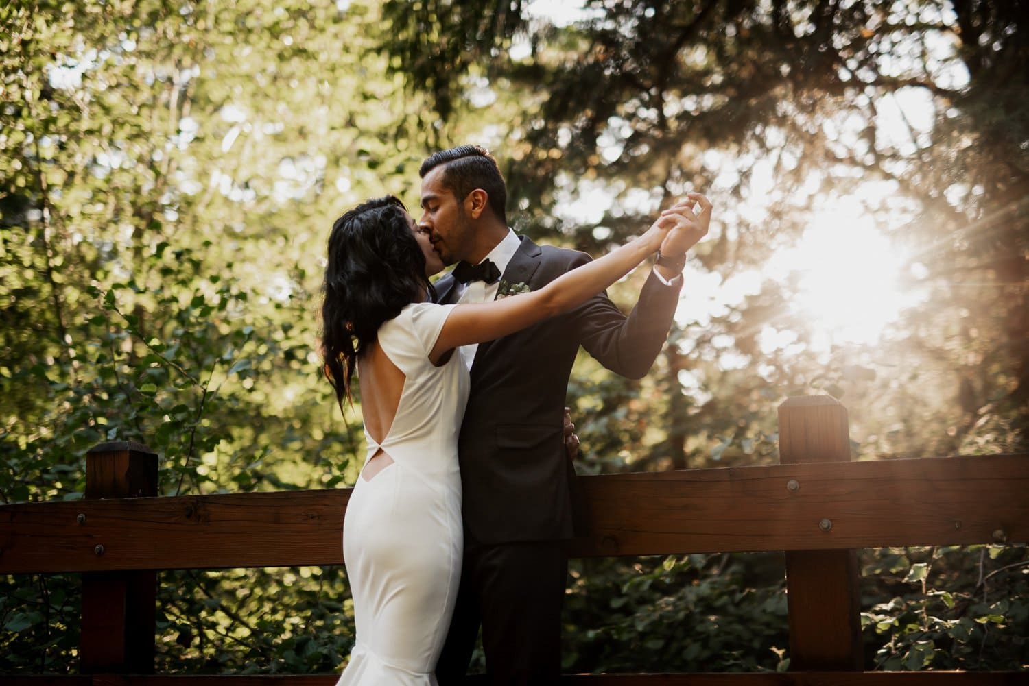 A couple dances on a wooden bridge at Leach Botanical Garden, with sunlight streaming through the trees and creating a magical wedding scene.