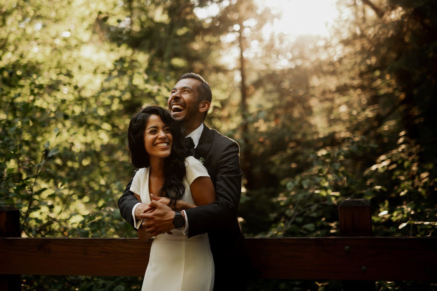 A couple in formal attire laughs and embraces on a wooden bridge surrounded by a sunlit forest at Leach Botanical Garden.